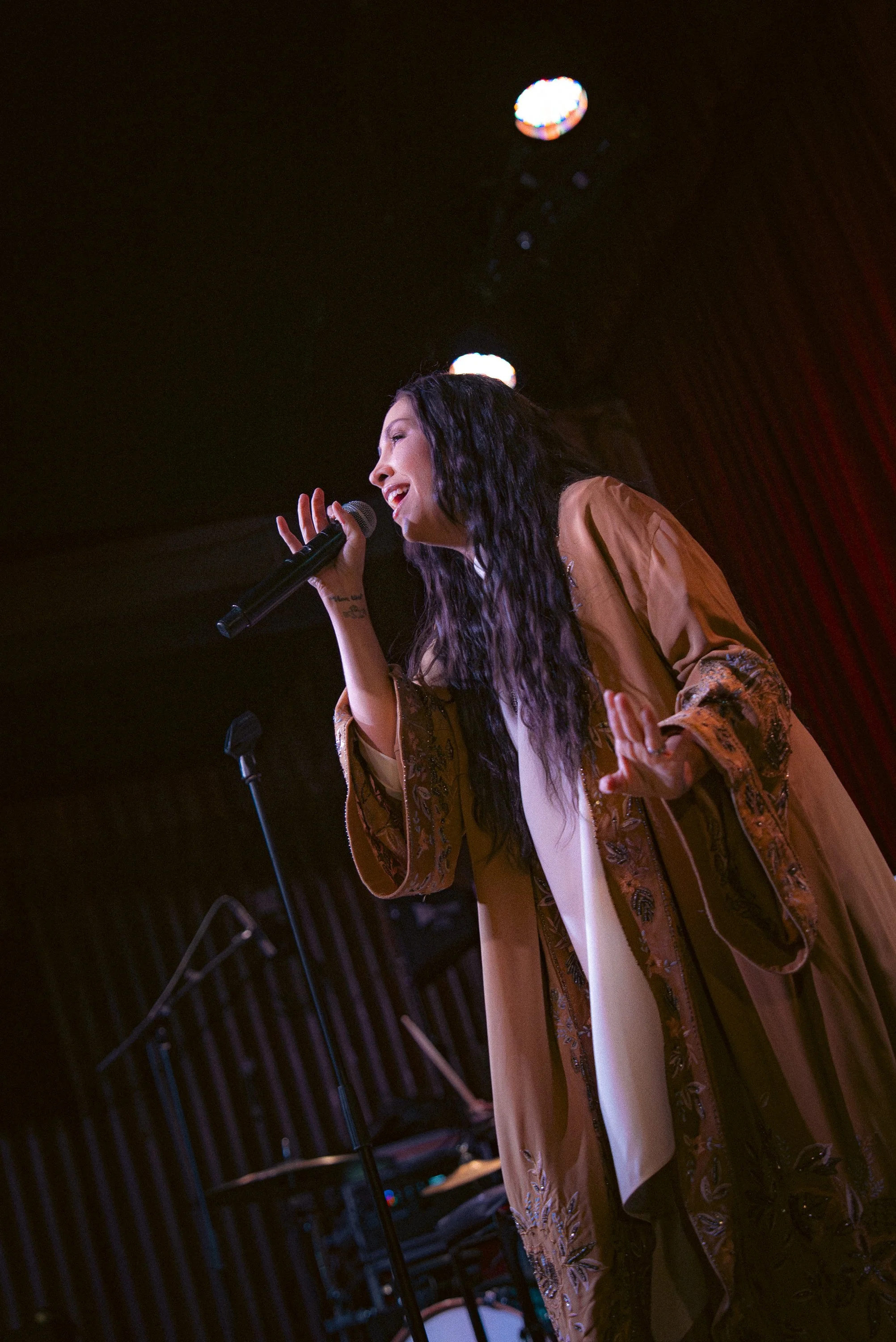 A woman with long, wavy black hair singing into a microphone on stage, smiling, wearing a tan and embroidered robe, with a dark background and stage lights.
