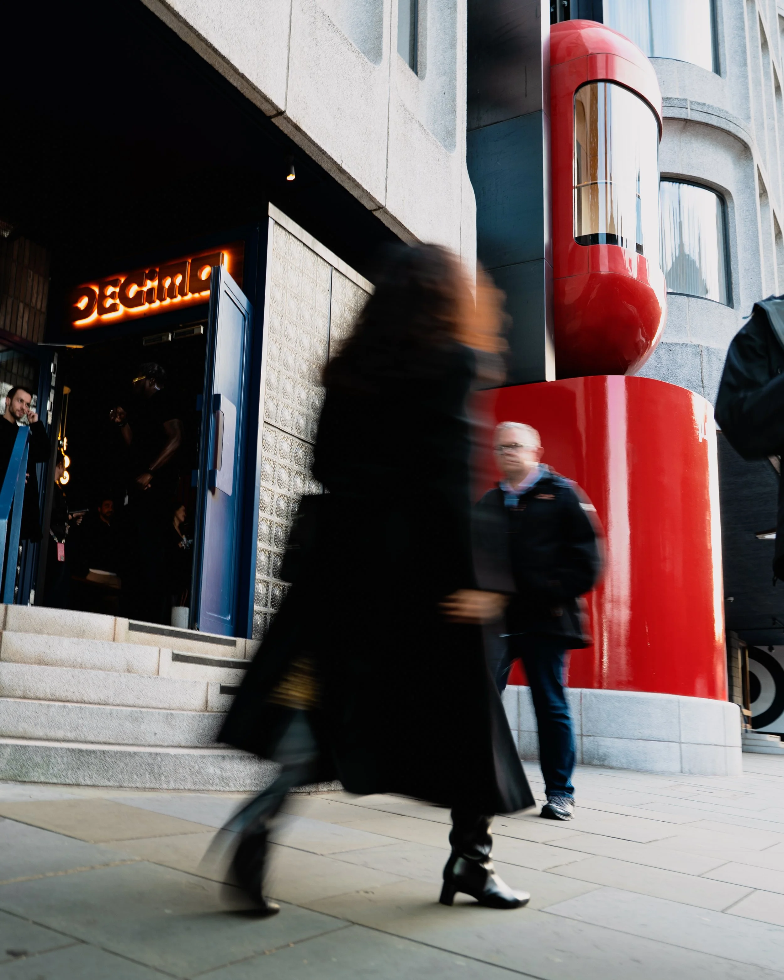 Blurred woman walking past the entrance of a restaurant or bar with a neon sign, with people inside and a large red cylindrical art piece outside on a city street.