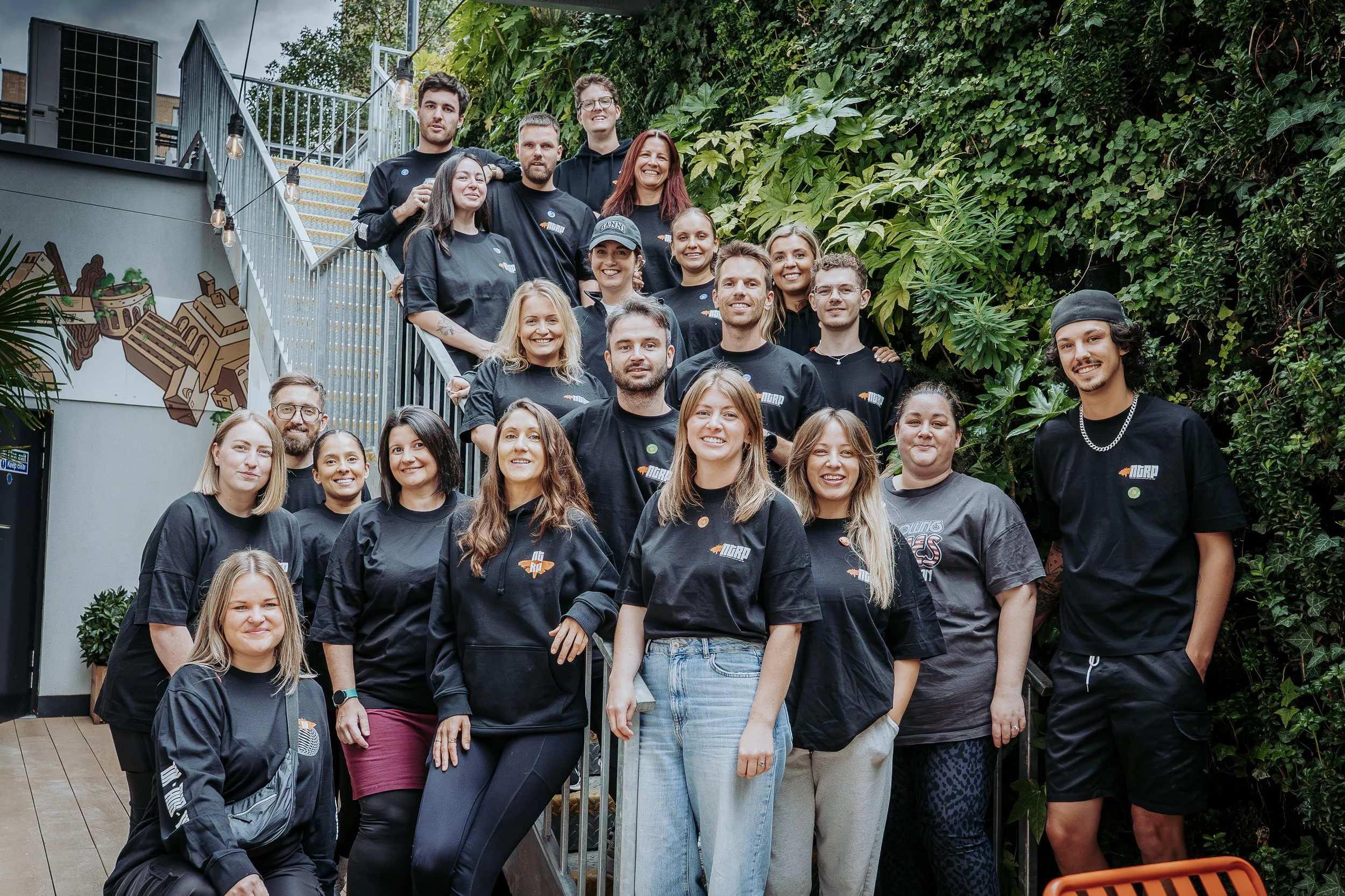 Group photo of smiling people standing and sitting on stairs and ground in front of a lush green wall, gathered outdoors for a team photo.