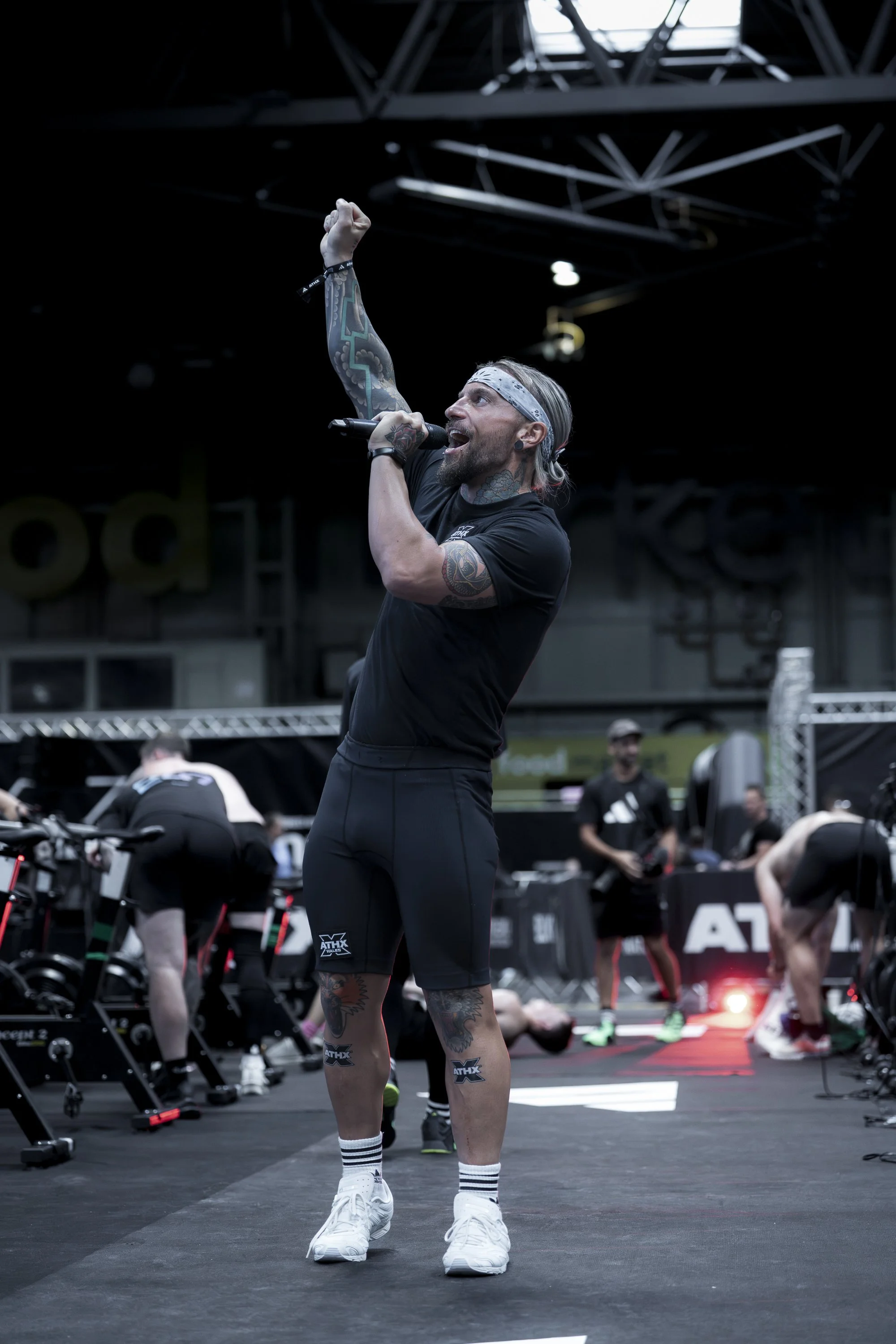 A man with tattoos, wearing a black shirt, black shorts, white sneakers, and a white headband, cheerfully speaking or singing into a microphone in a gym or fitness event setting.