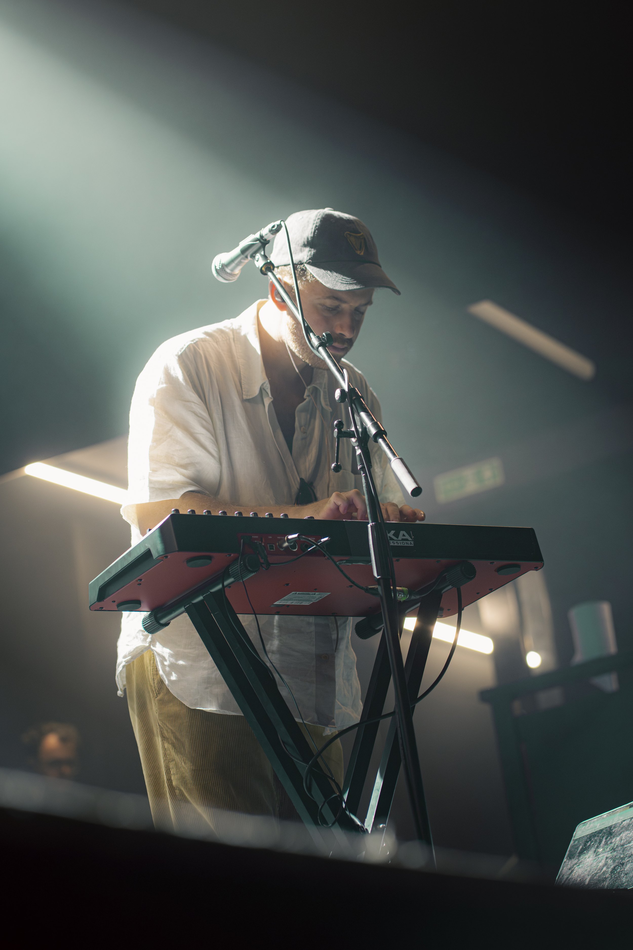 A man wearing a baseball cap and light-colored shirt playing a keyboard on stage, with a microphone in front of him.