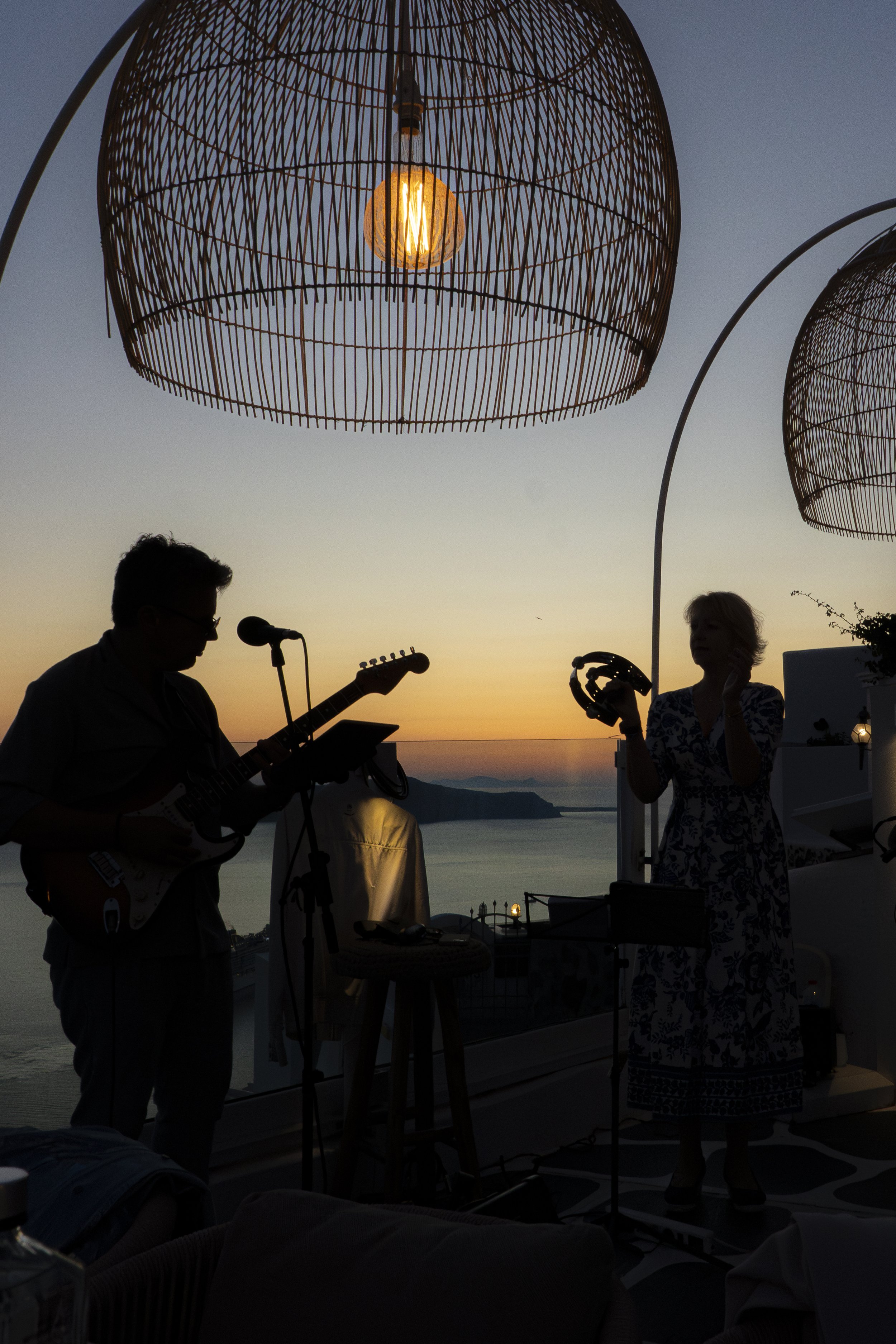 Silhouettes of two people, one playing guitar and the other holding a tambourine, during sunset, with large woven light fixtures hanging above.