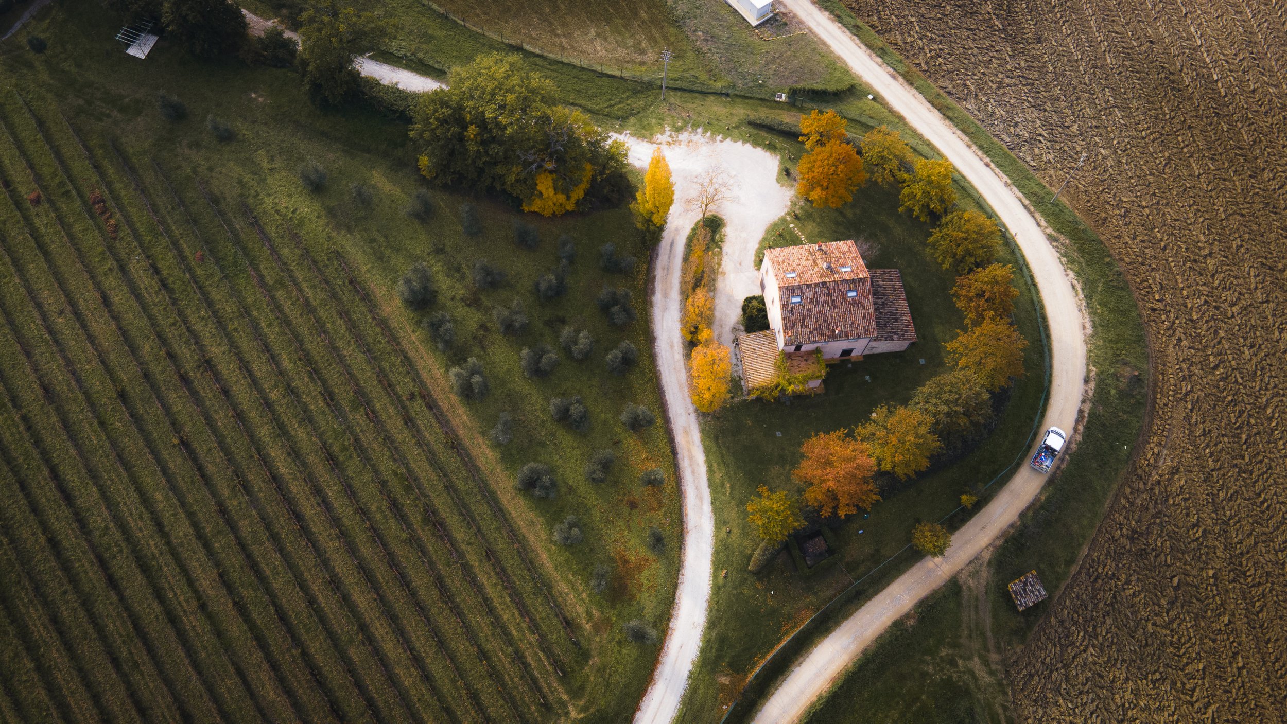 An aerial view of a rural farmhouse surrounded by trees with colorful autumn foliage, a gravel driveway, adjacent farmland, and a white vehicle parked on the driveway.