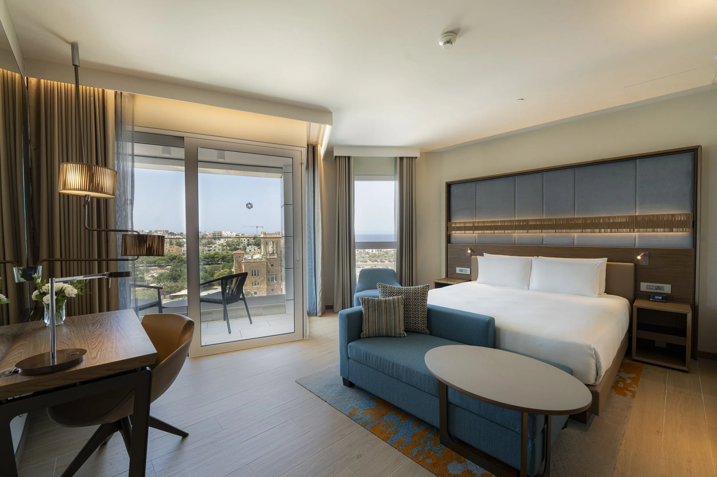 Modern hotel room featuring a large bed with white linens, a blue sofa with decorative pillows, a wooden desk with a lamp, balcony with outdoor chairs, and large windows with a city view.