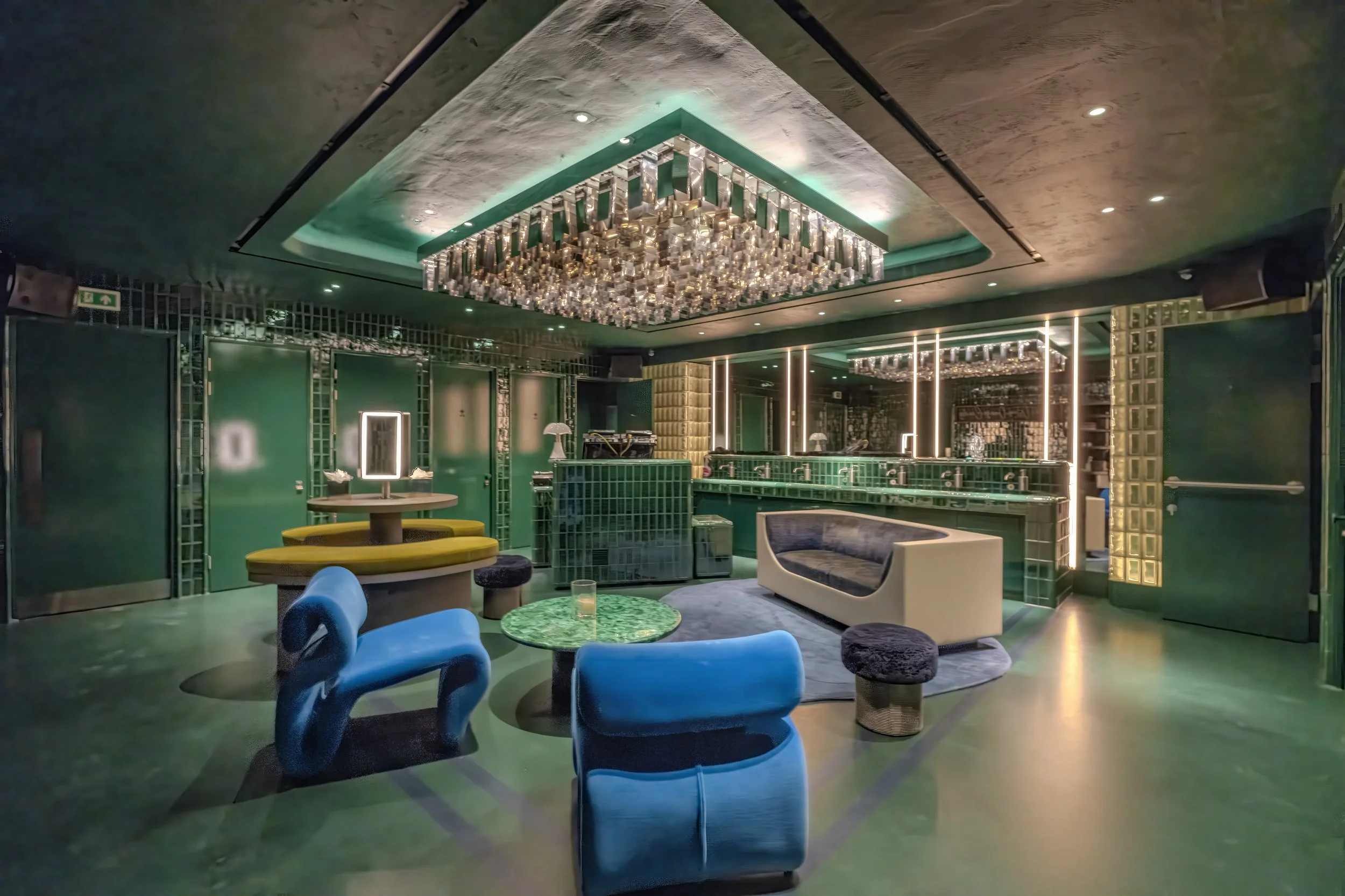 Interior of a stylish lounge bar with green tile accents, modern furniture, blue and black chairs, round tables, and a chandelier ceiling light.
