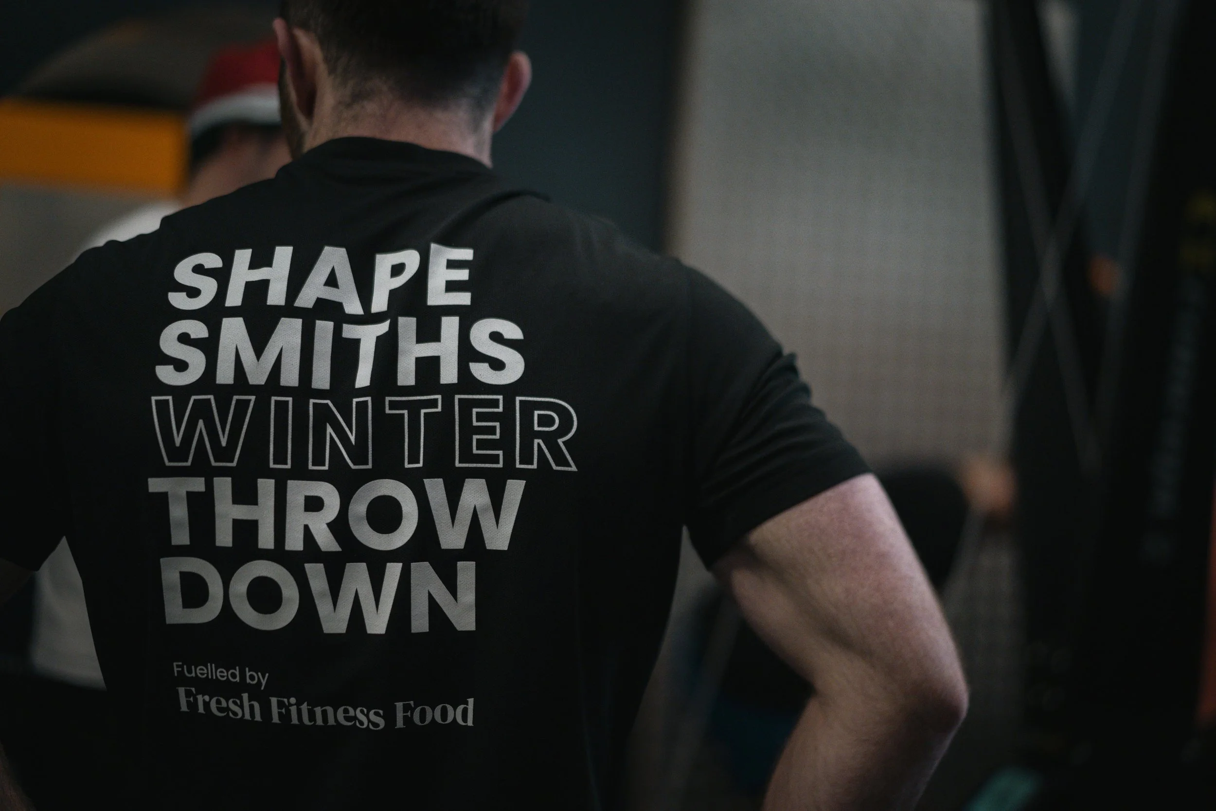Back of a man wearing a black T-shirt with bold white and outlined text that reads, "Shape Smiths Winter Throw Down." The shirt also has smaller text that says, "Fuelled by Fresh Fitness Food." The setting appears to be a gym or fitness event.