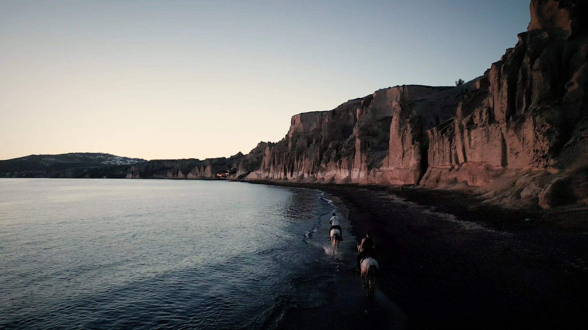People riding horses along a dark beach at sunset with cliffs in the background.