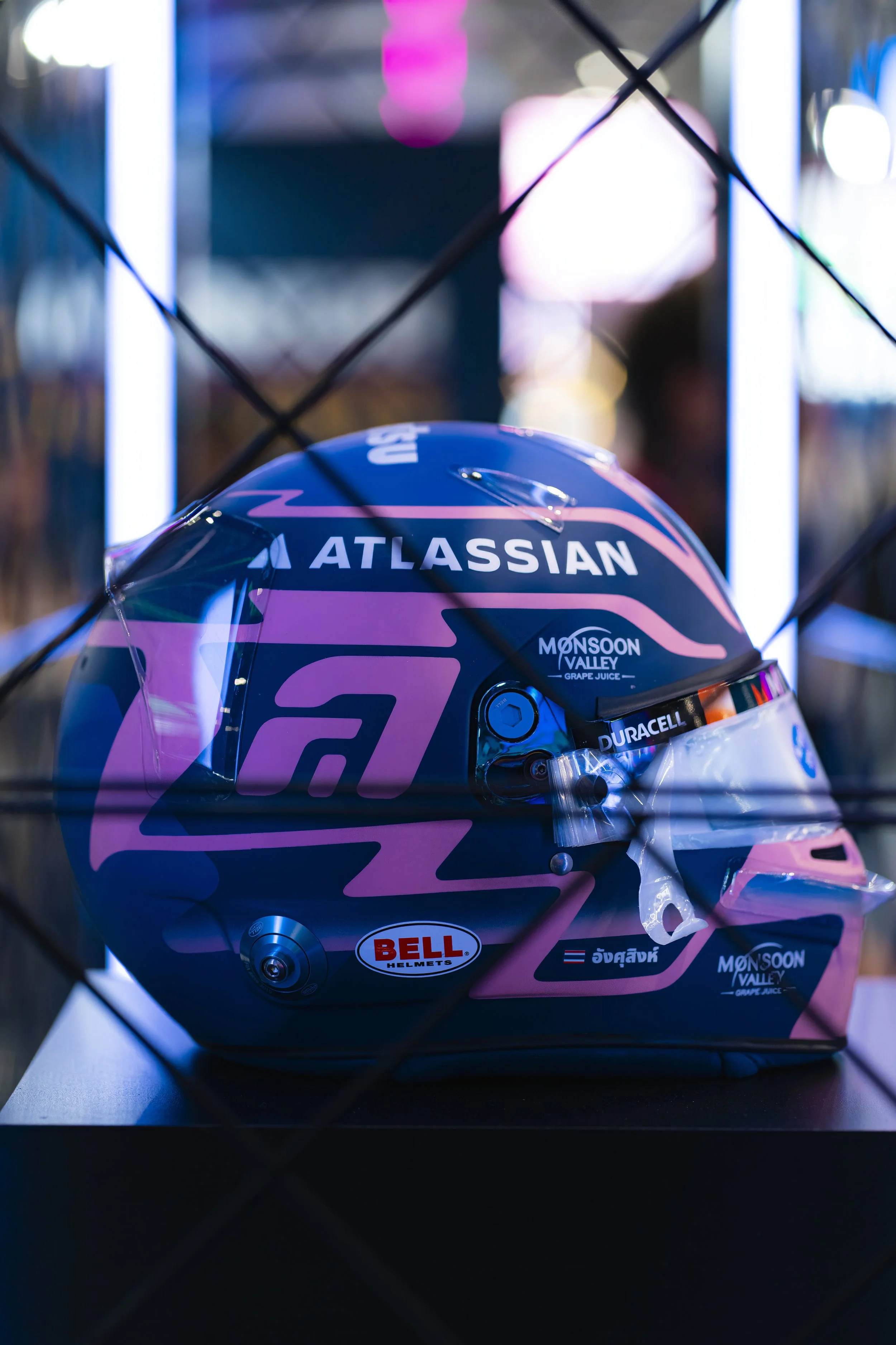 A racing helmet with a dark blue and pink design, featuring sponsorship logos such as Atlassian, Bell Helments, Duracell, and Monsoon Valley Grape Juice, displayed behind a wire mesh.