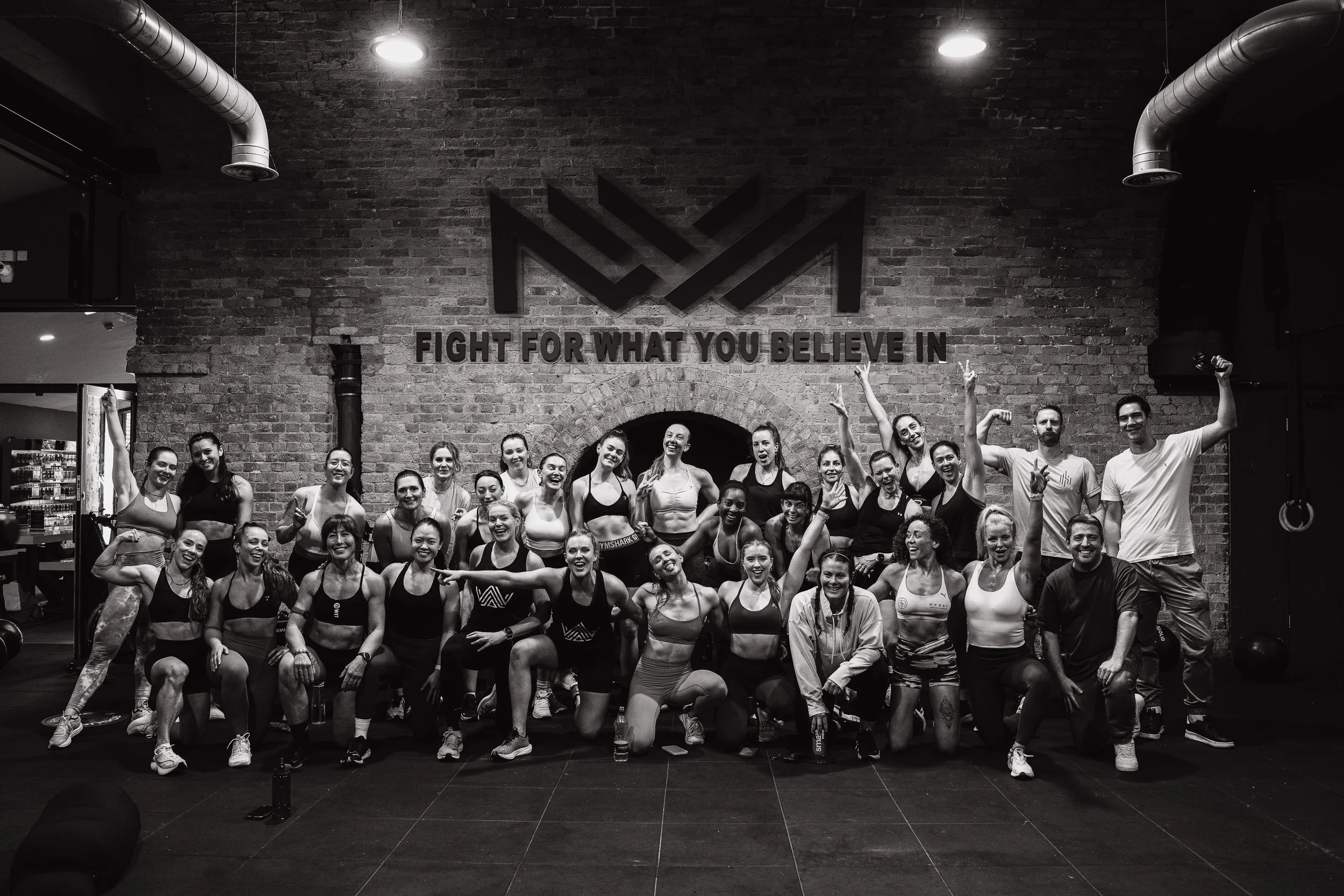 A group of women and a few men in workout gear posing together in a gym with a brick wall and the phrase 'Fight for What You Believe In' on the wall behind them, some lifting their arms in victory.