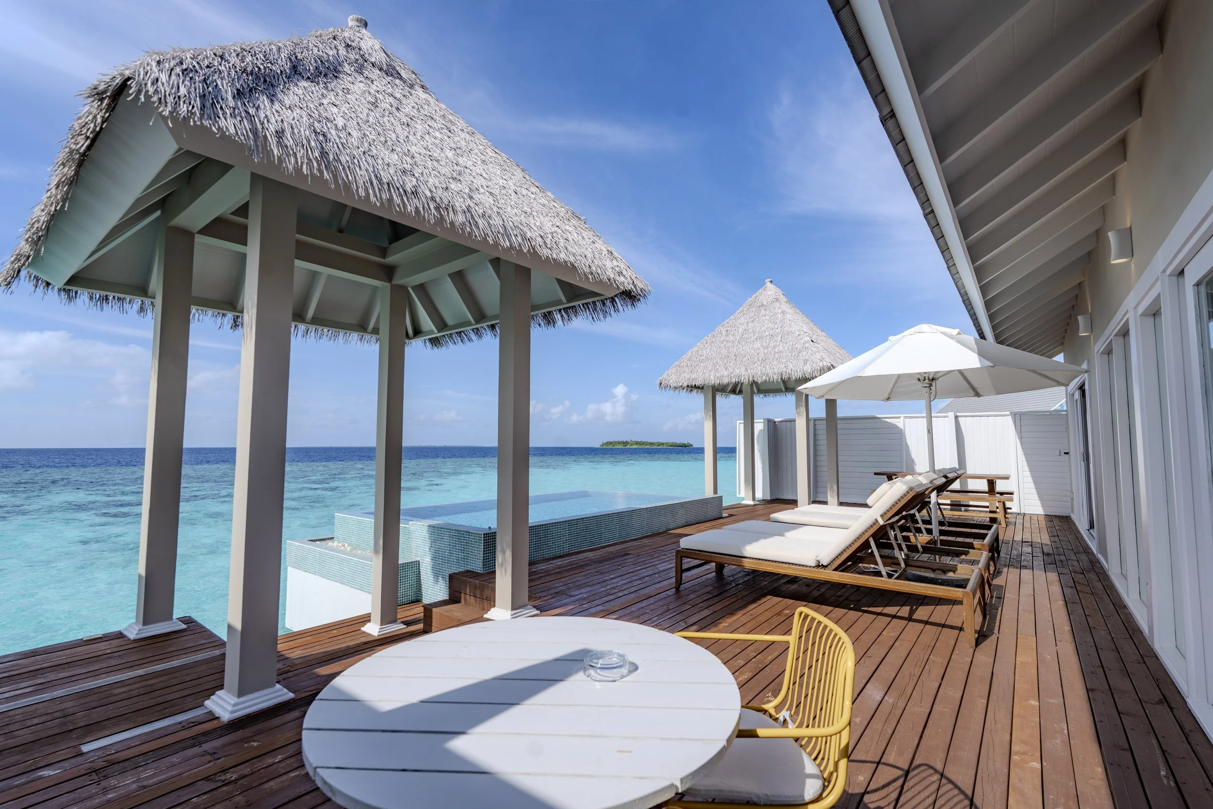 Beachside deck with thatched-roof cabanas, lounge chairs, white umbrellas, blue ocean, and clear sky.