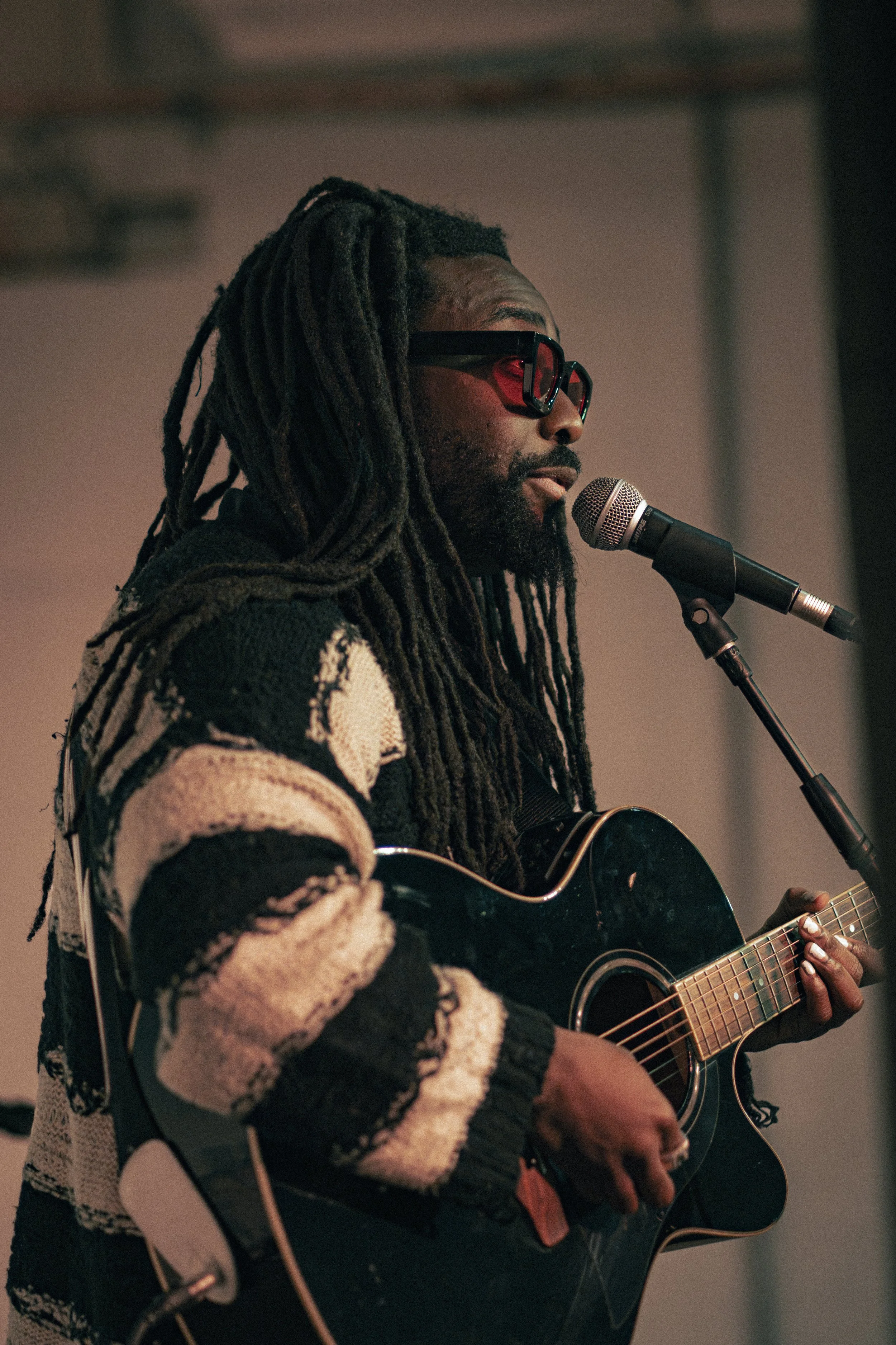 A man with long dreadlocks, wearing sunglasses and a patterned sweater, plays an acoustic guitar and sings into a microphone.