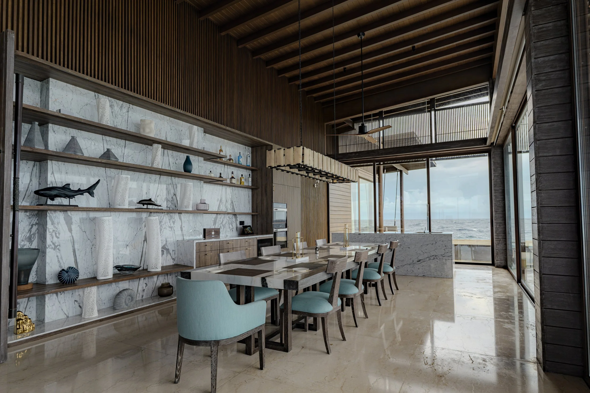 Modern seaside dining room with a marble dining table, turquoise chairs, and a large window overlooking the ocean.