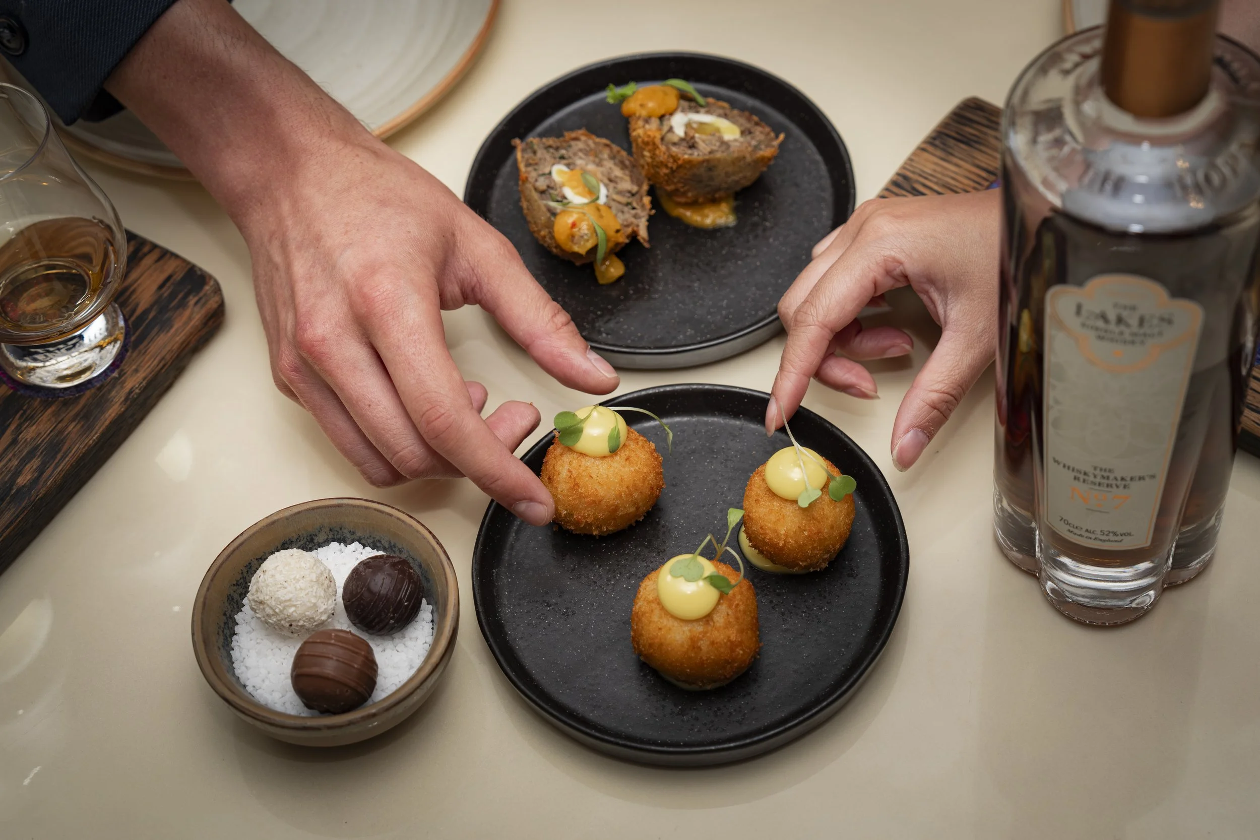 Hands reaching for three golden-brown croquettes topped with white sauce and microgreens, placed on black plates, with a small bowl of assorted chocolates, a glass of whiskey, a bottle of spirits, and a dish of pâté or terrine in the background.
