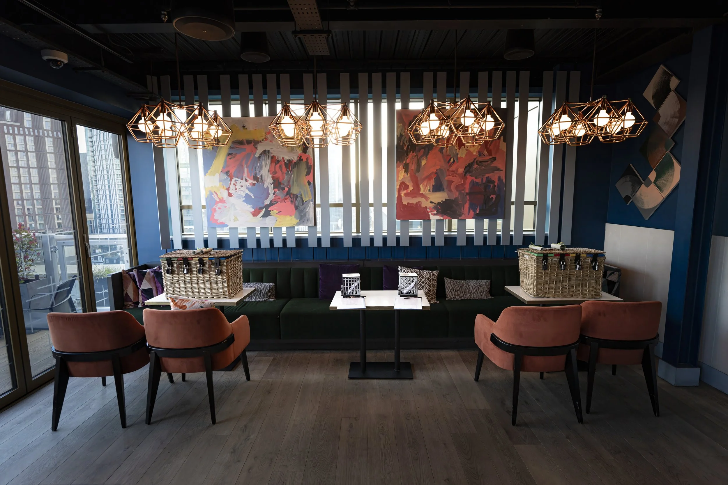 Interior of a modern restaurant or cafe with a dark ceiling, orange pendant lights, blue wall with colorful abstract paintings, and a long green booth with pillows. There are four pink chairs in front of the booth and small tables with wicker baskets