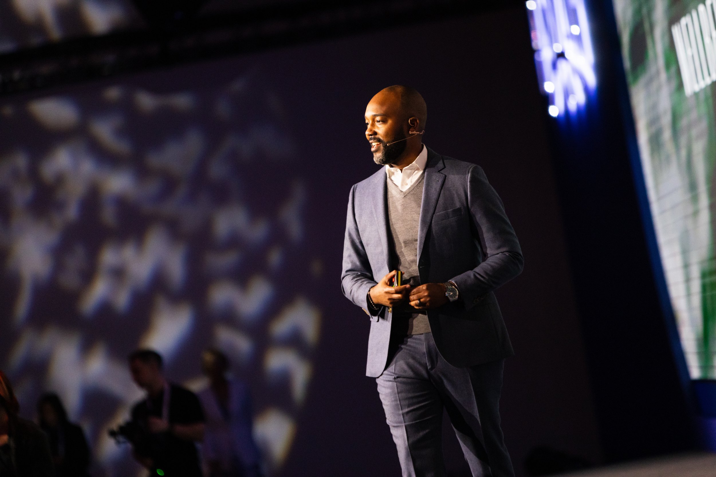 A man in a dark gray suit giving a presentation on stage, holding a remote, with a blurred audience in the background.