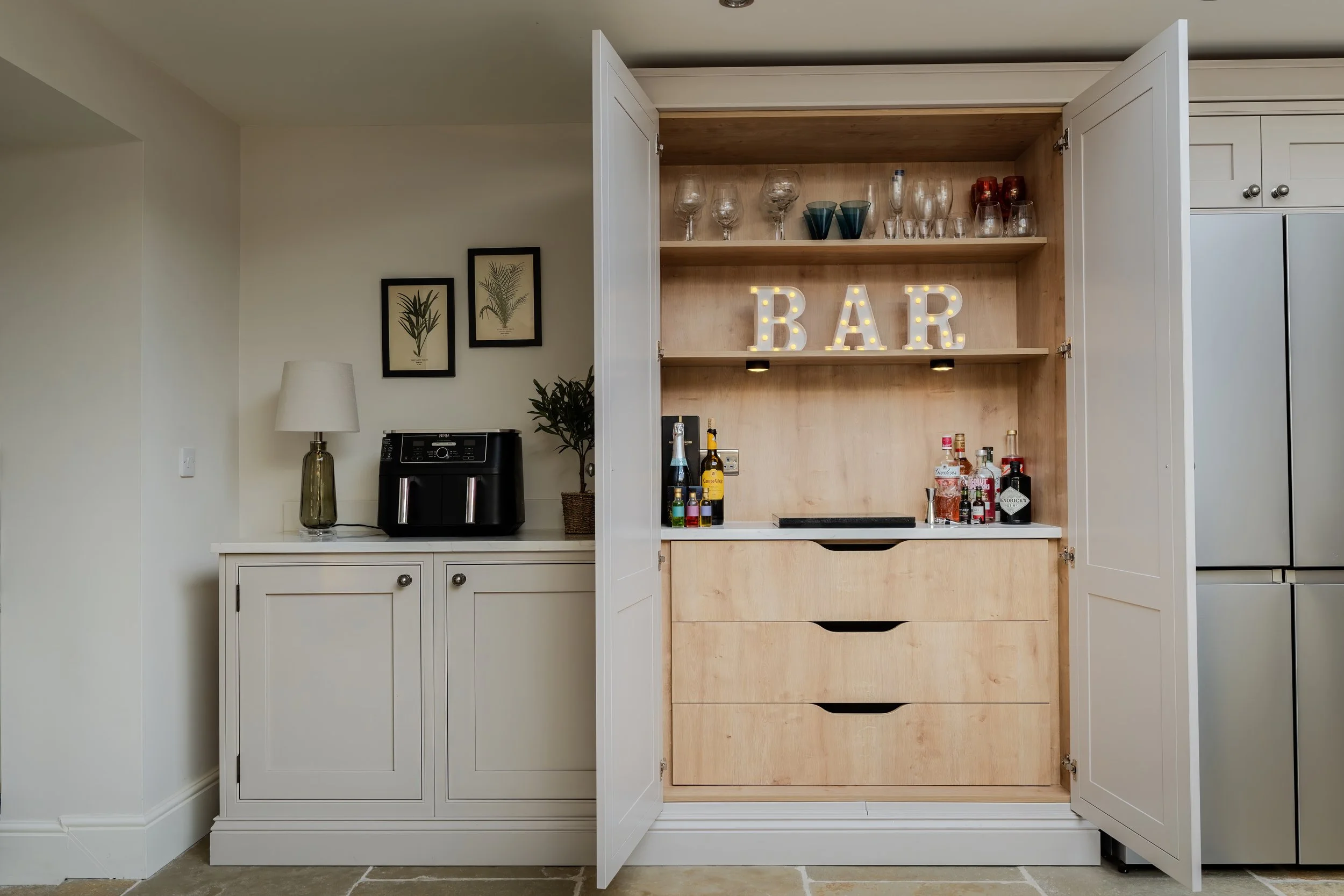 Open kitchen cabinet with bar setup, including liquor bottles, glassware, and illuminated 'BAR' sign, adjacent to a white cabinet with a lamp, plant, and air fryer.