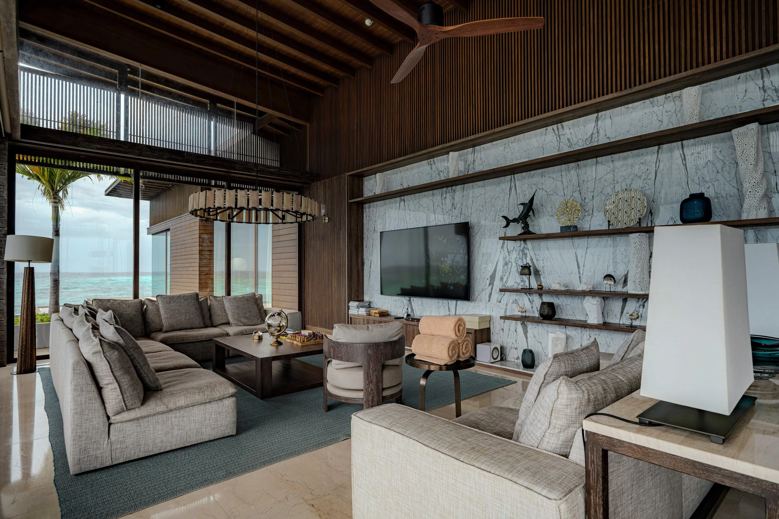 Modern living room with large sectional sofa, armchair, marble wall, wall-mounted TV, shelves with decorative items, and ocean view outside.