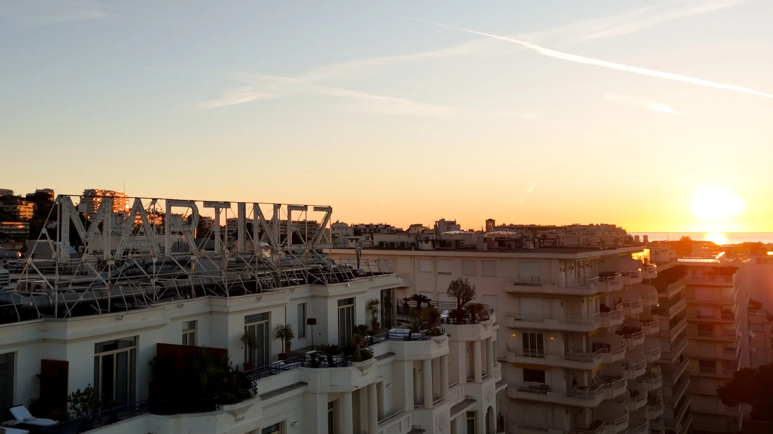 Sunset over a city with modern apartment buildings and a rooftop with some plants, with the sea in the background and the sun near the horizon.