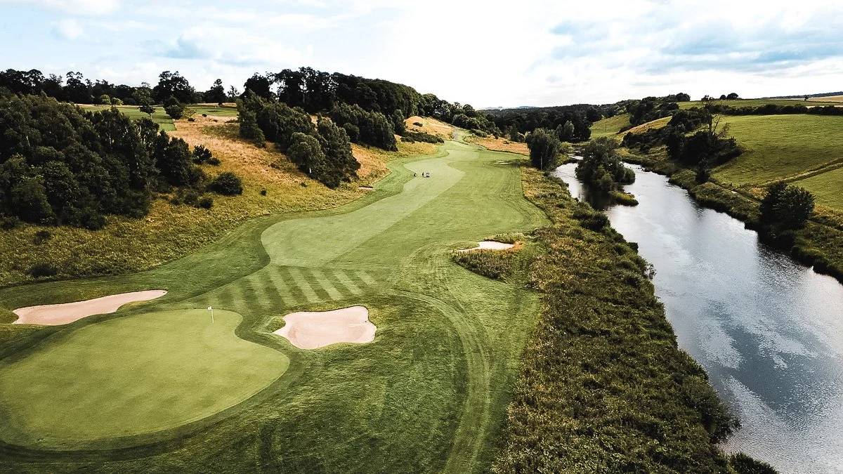 Aerial view of a golf course with sand bunkers, green fairways, and a nearby river with trees lining its banks.