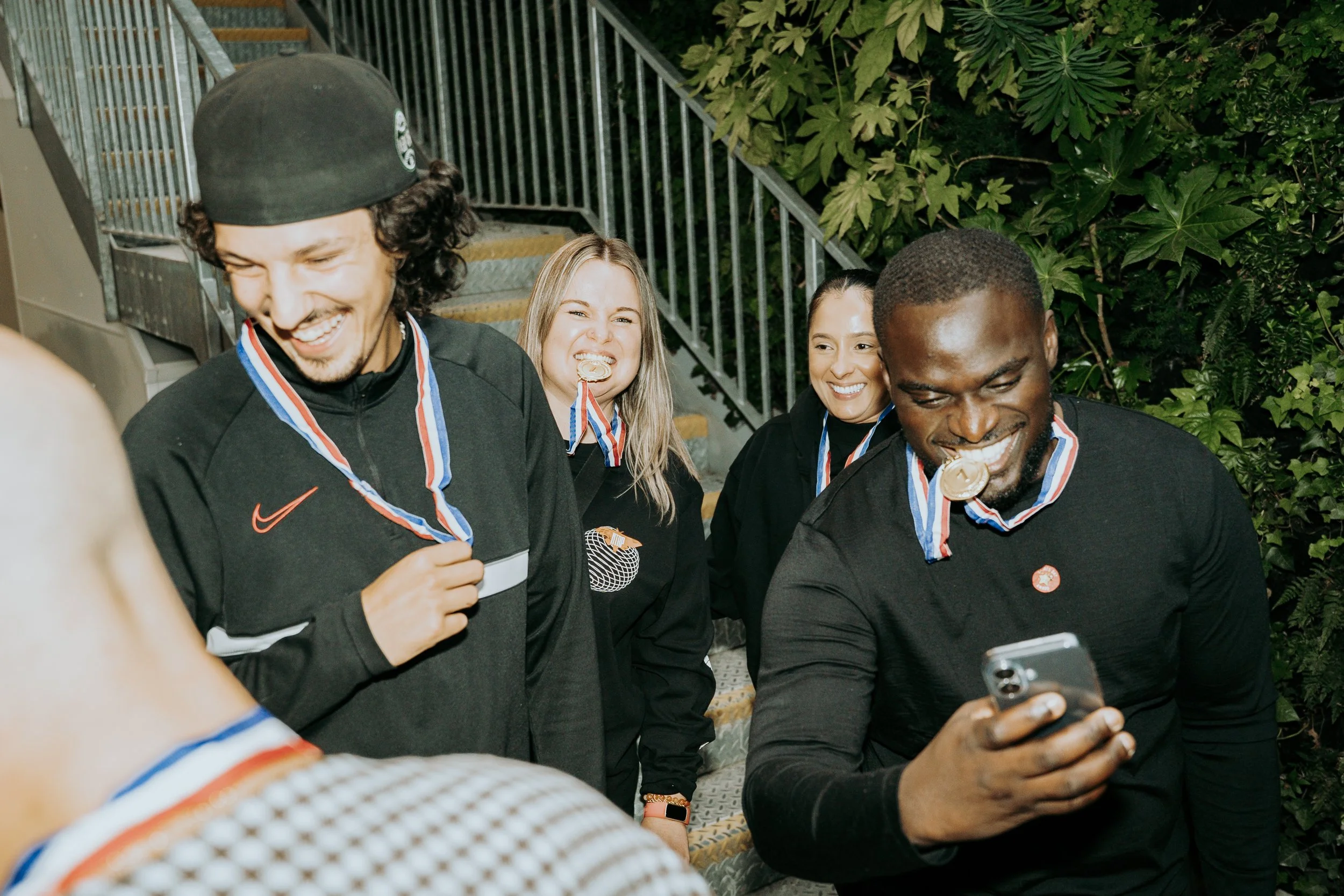 Four athletes with medals around their necks celebrating and smiling, standing on stairs near green foliage.