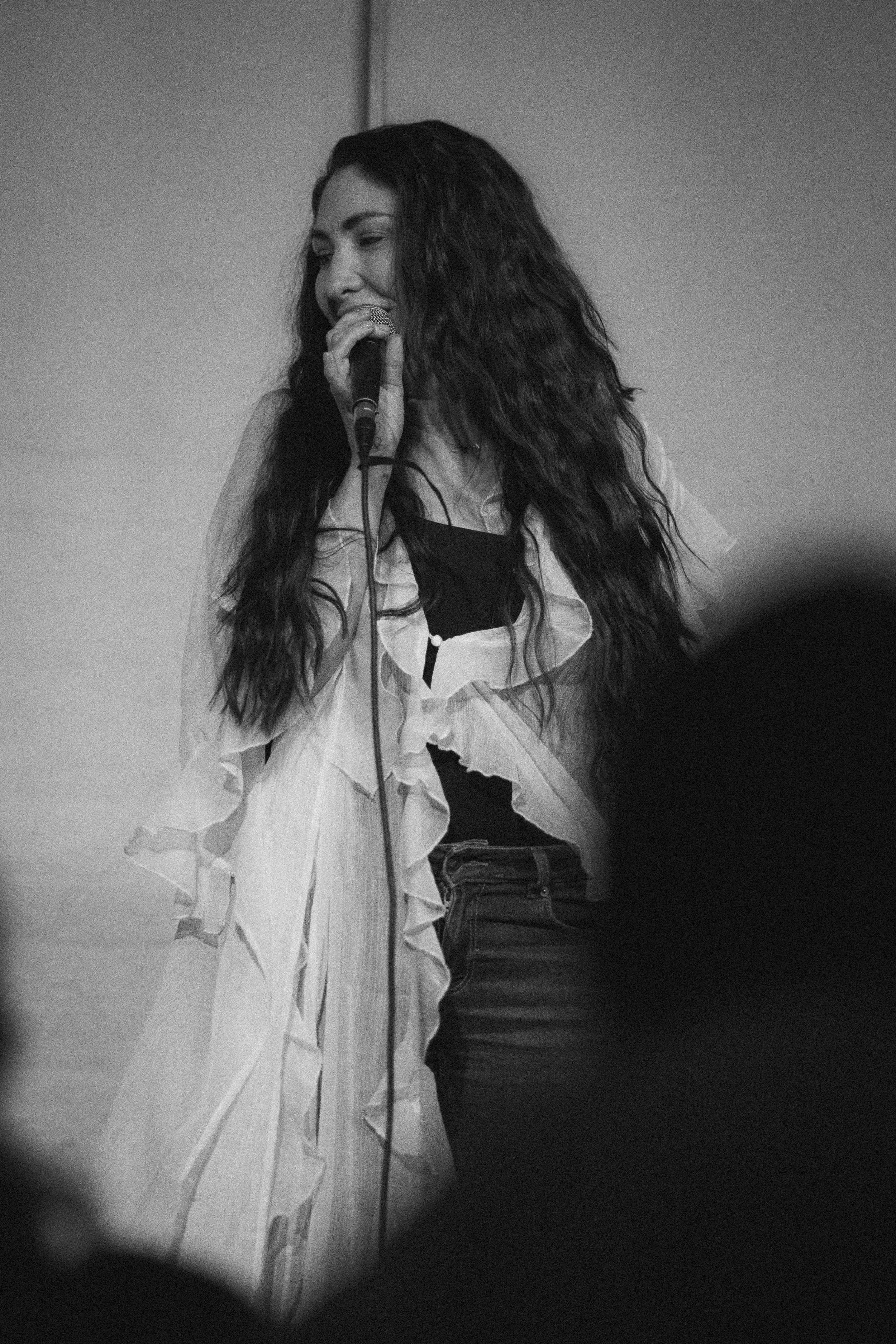 A woman with long, curly hair singing into a microphone on stage, with blurred audience in the foreground.