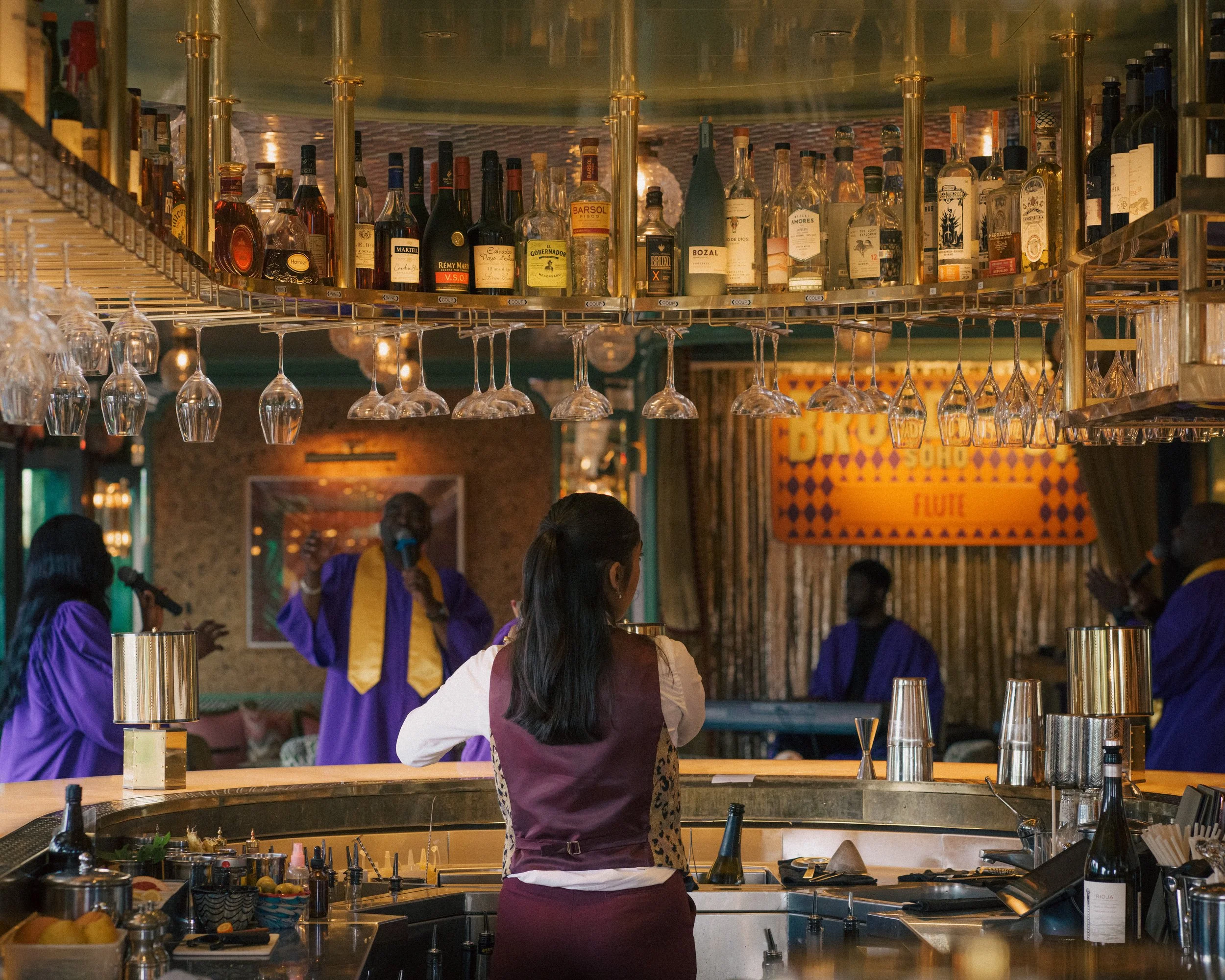 A bartender preparing drinks at a bar, with several bottles of alcohol and hanging wine glasses overhead, while four patrons in purple attire are engaged in conversation at the bar.