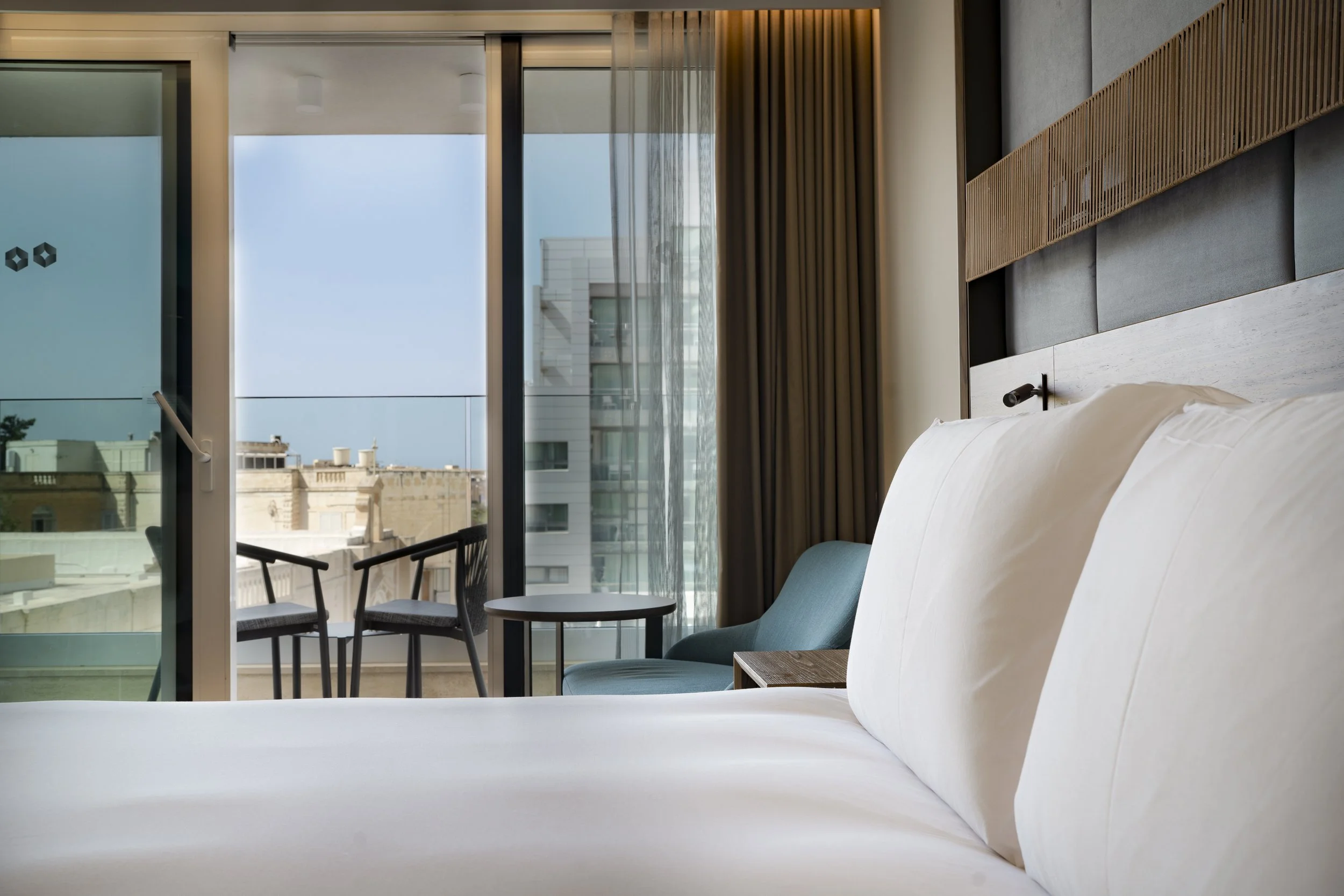 A hotel room with a bed, a balcony with two chairs and a table, with city buildings and the sky outside.