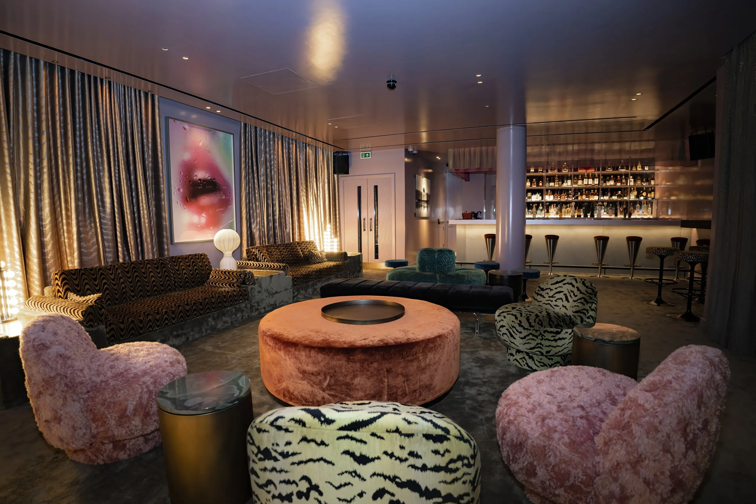 Luxurious lounge with patterned velvet chairs, a large pink circular sofa, a bar in the background with illuminated shelves of liquor bottles, a tall white pillar, framed artwork of a face with red lips and water droplets, and warm ambient lighting.