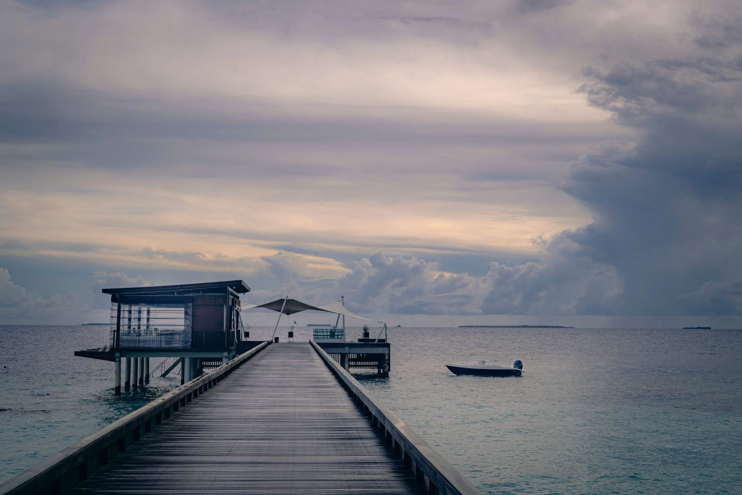 A wooden pier extending over calm water leads to a small building with a slanted roof and large windows, with a boat nearby, under a cloudy sky.