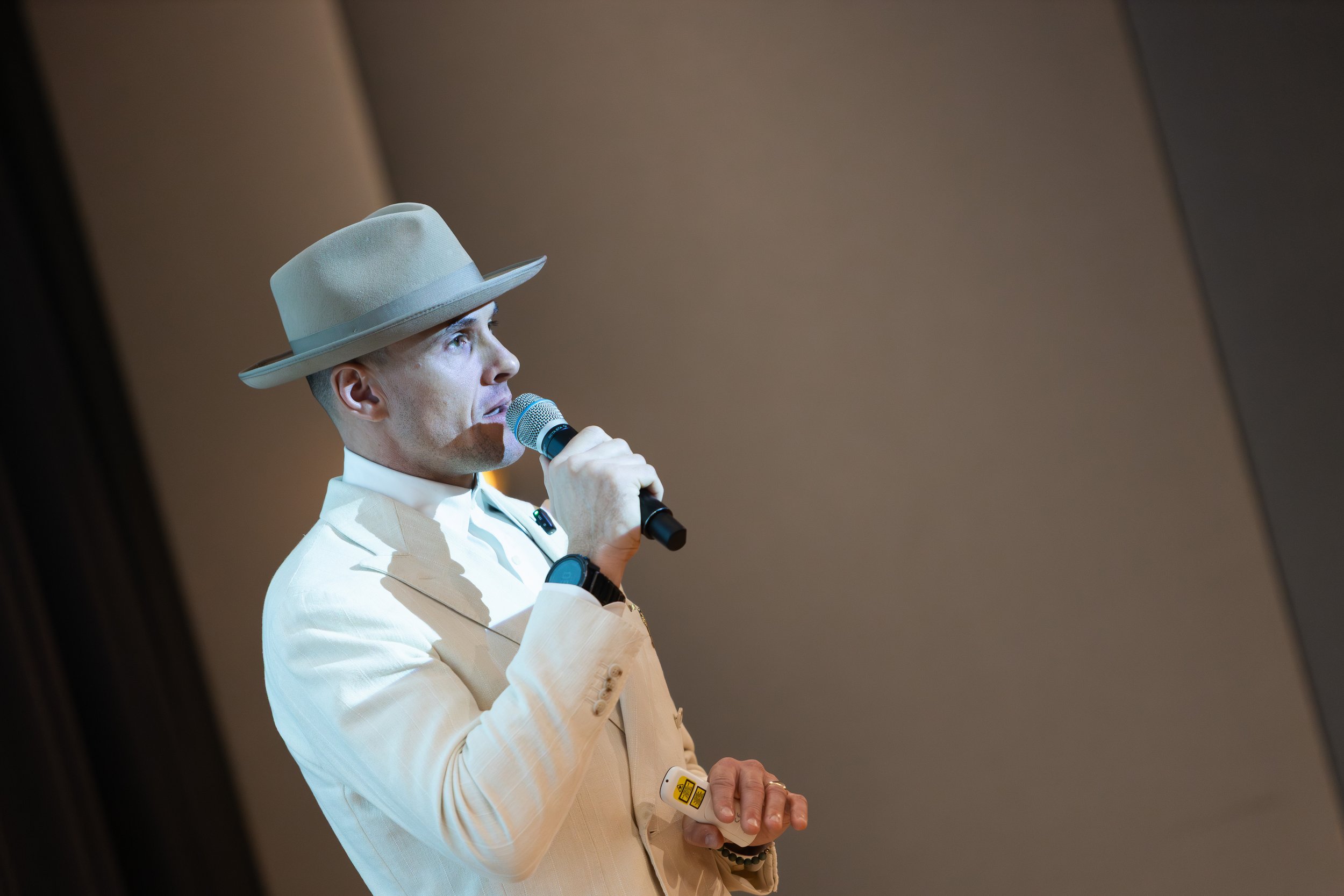 Man in a beige suit and fedora hat speaking into a microphone on stage.