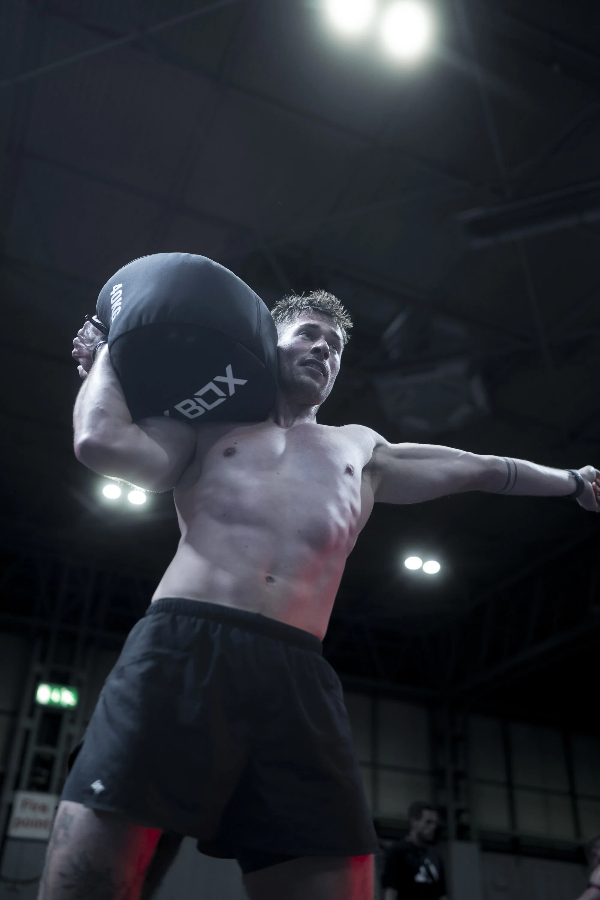 A shirtless man participating in a fitness activity, holding a large weighted bag on his shoulder, inside a gym with bright lights overhead.