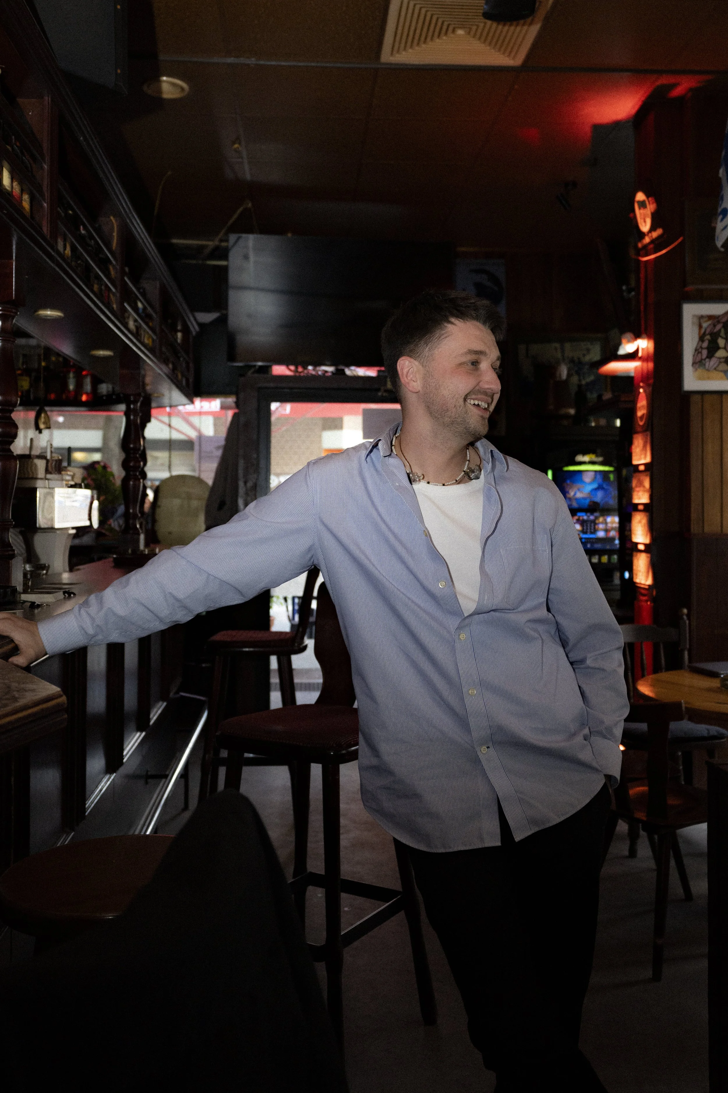 A smiling man in a light blue shirt and white t-shirt standing inside a bar or restaurant, leaning against the bar with a wooden shelf filled with bottles in the background.