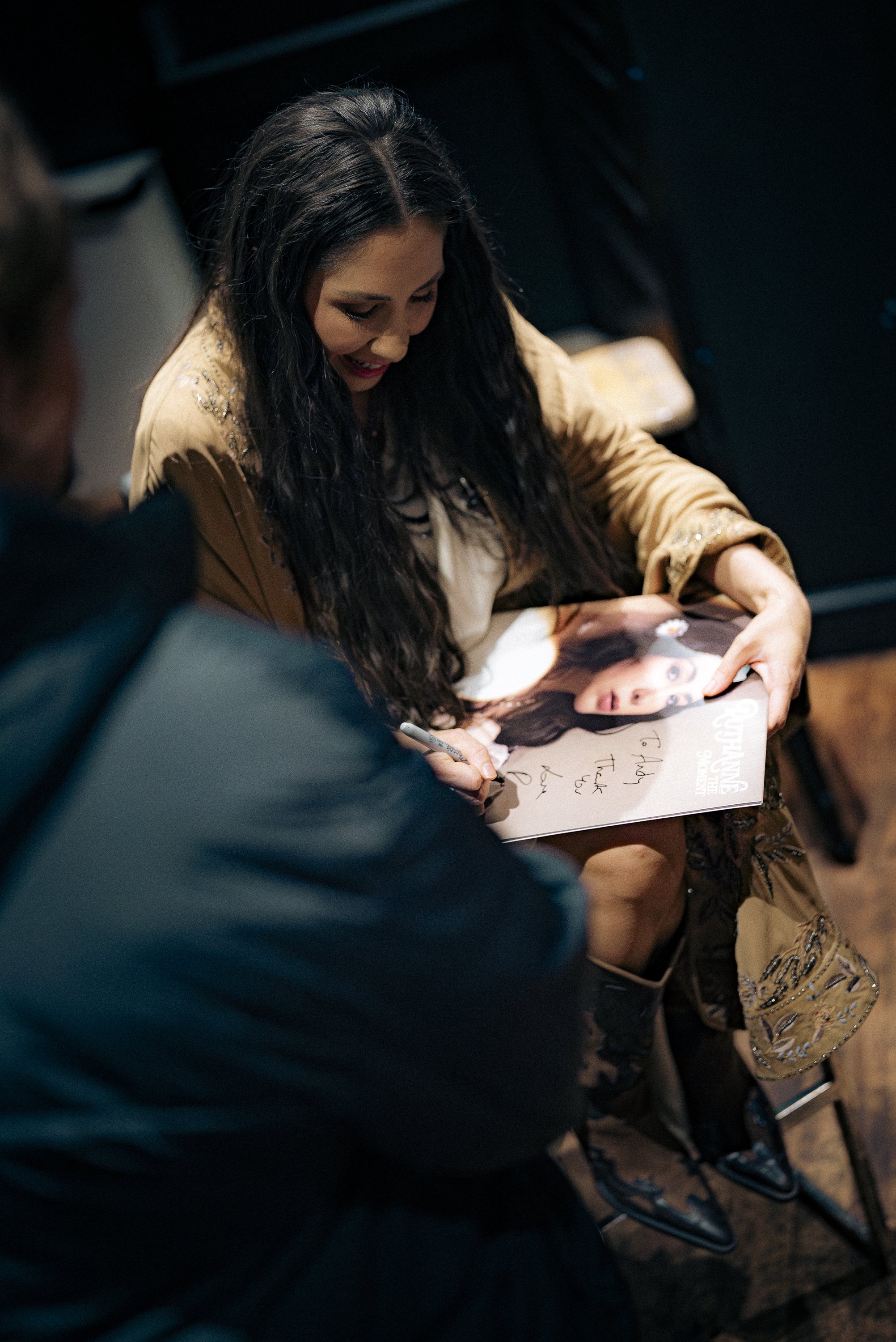 A woman with long dark hair signing an autograph for a fan holding a magazine with a photo of her on the cover in an indoor setting.