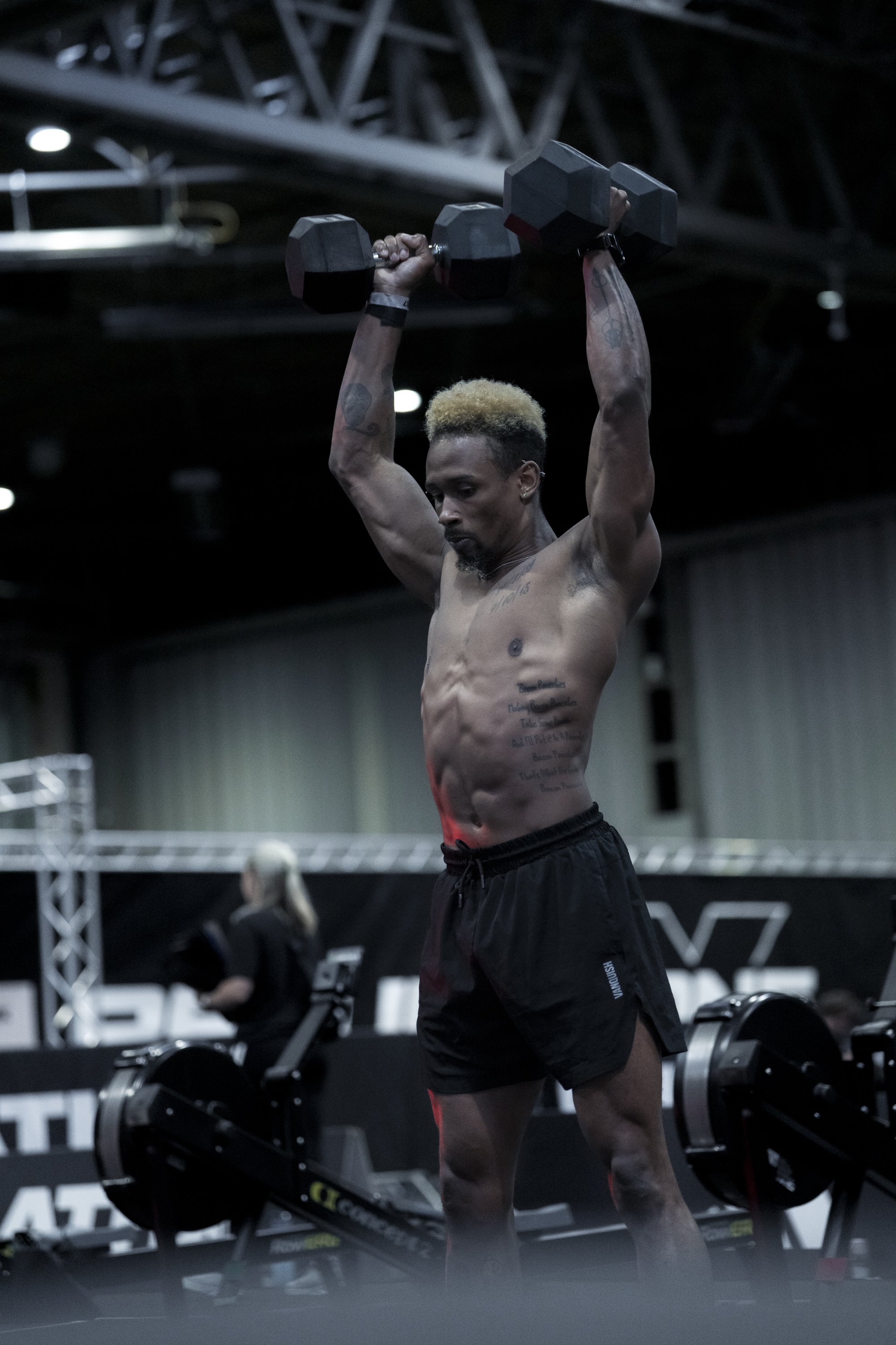 A shirtless man with tattoos exercises with a dumbbell in a gym, standing in front of rowing machines.
