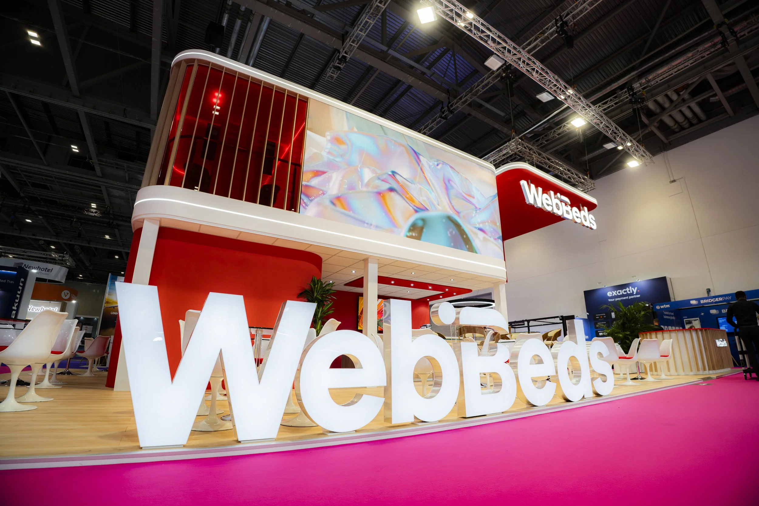 Trade show booth for WebBeds featuring a large illuminated sign spelling 'WebBeds' at the front, a two-story structure with a large digital screen displaying colorful abstract visuals, seating area with white chairs, and decorative plants inside an e