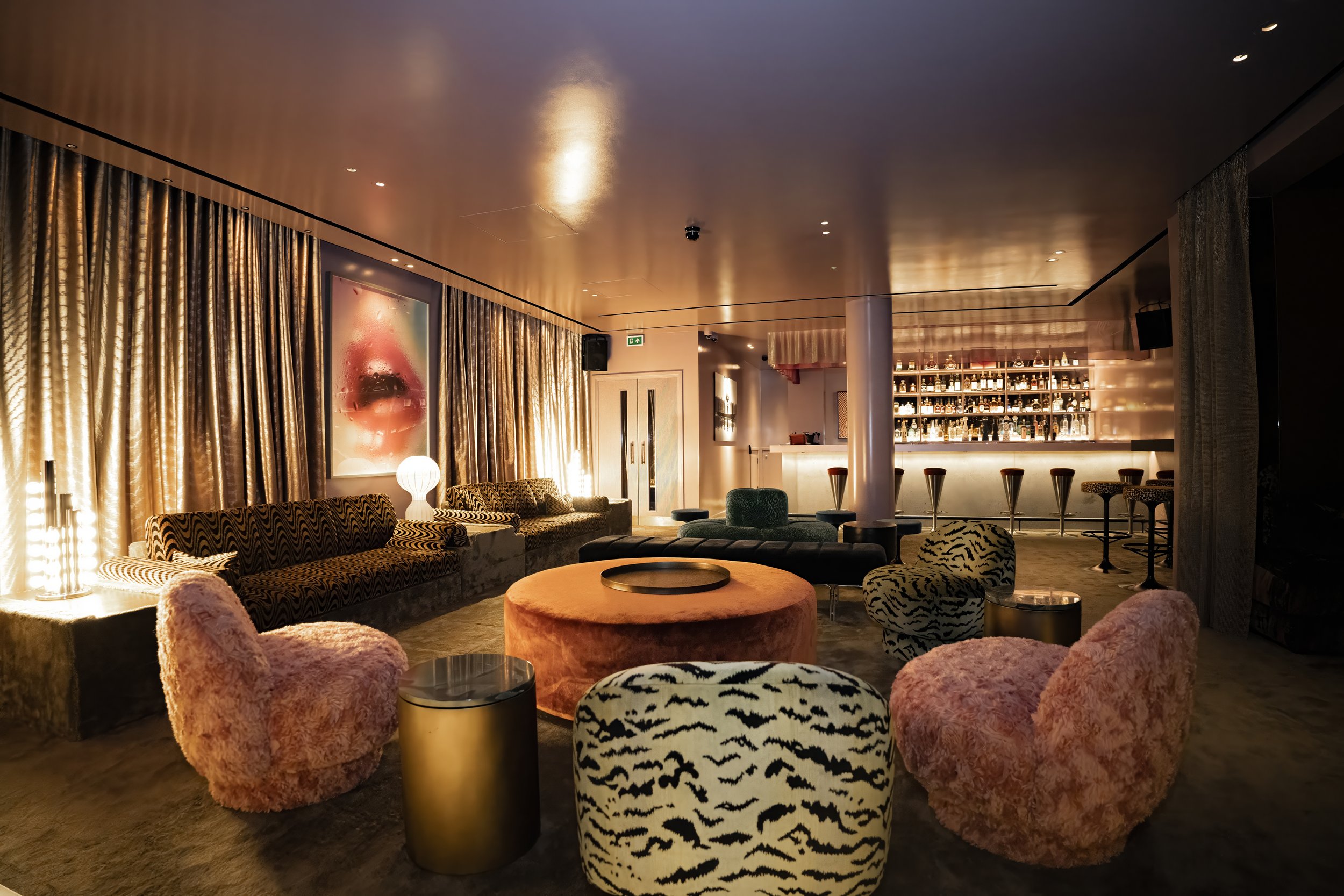 Luxurious lounge with velvet and plush chairs, bar area with bottles on shelves, golden curtains, and artwork on the wall.