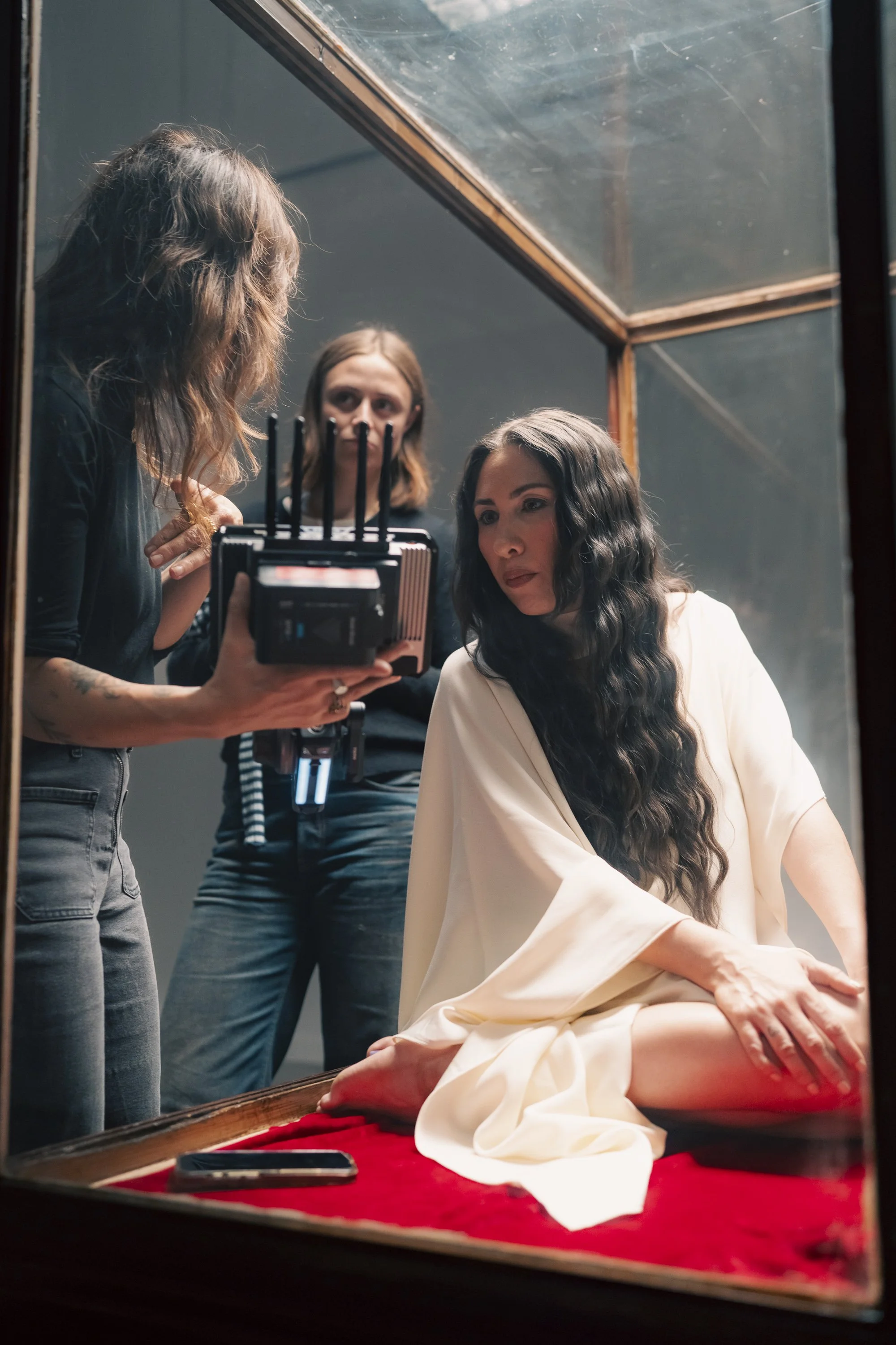 Woman with long wavy black hair sitting on a red velvet cushion, wearing a white robe, being filmed by two women in a dimly lit room with a mirror.