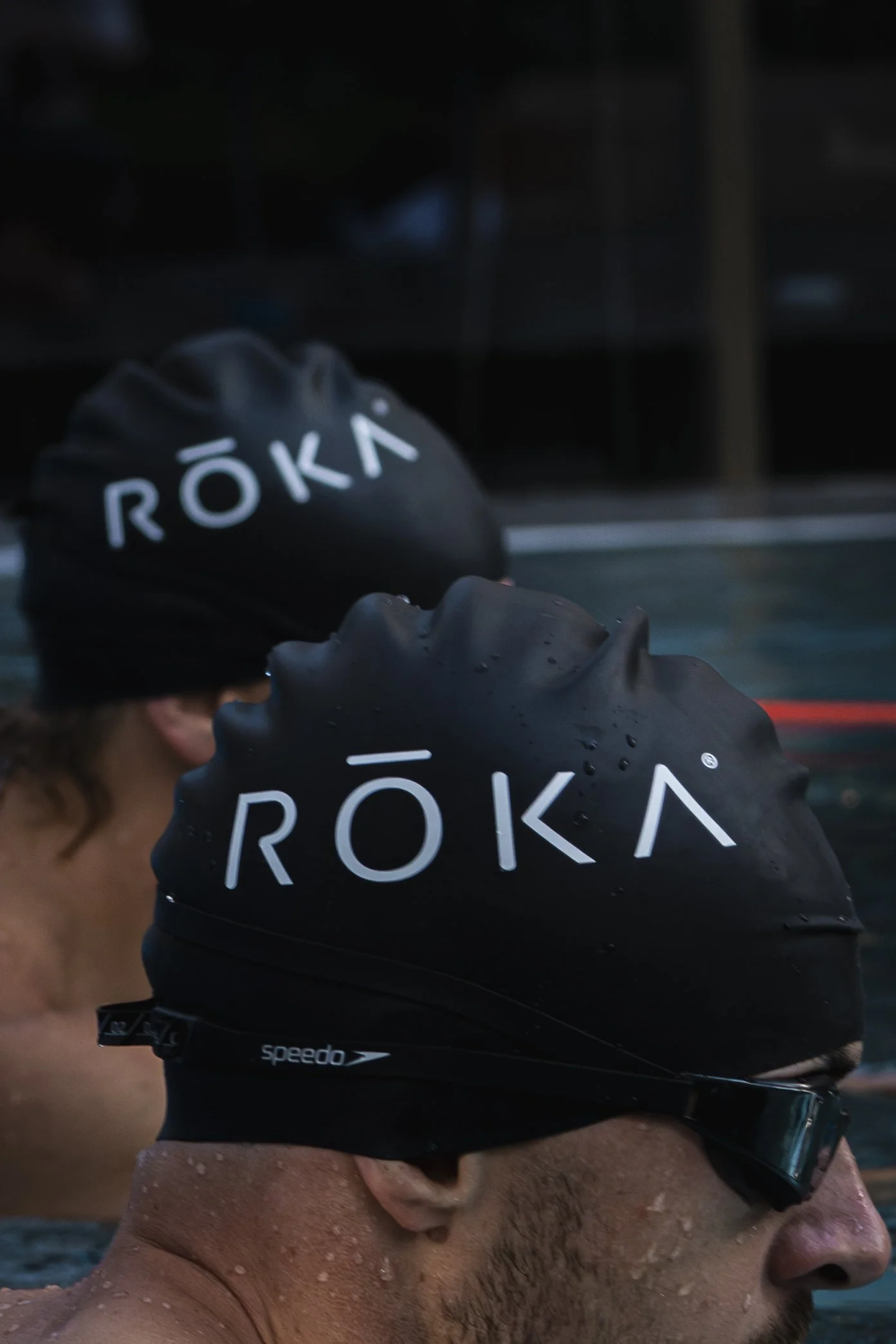 Two swimmers wearing black swimming caps with the word 'RŌKA' printed on them, preparing for a race.