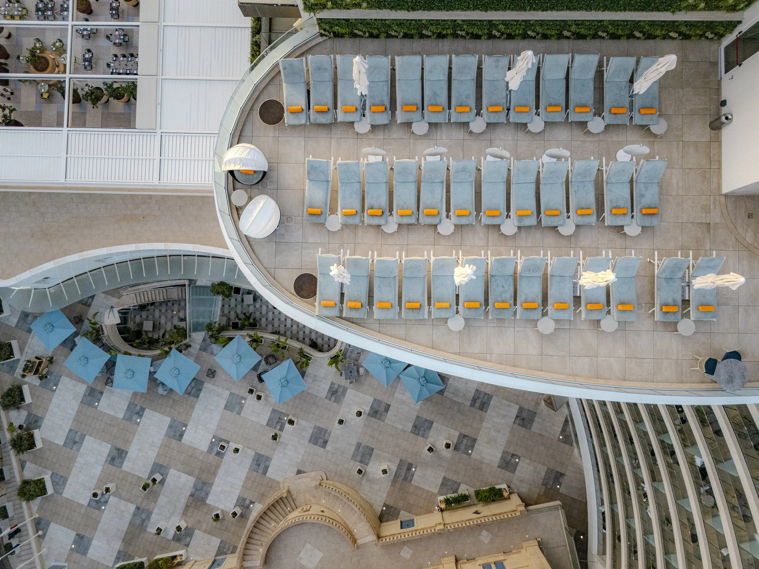 Aerial view of a rooftop outdoor area with rows of blue lounge chairs, umbrellas, and small tables, overlooking a lower level with blue umbrellas and potted plants.
