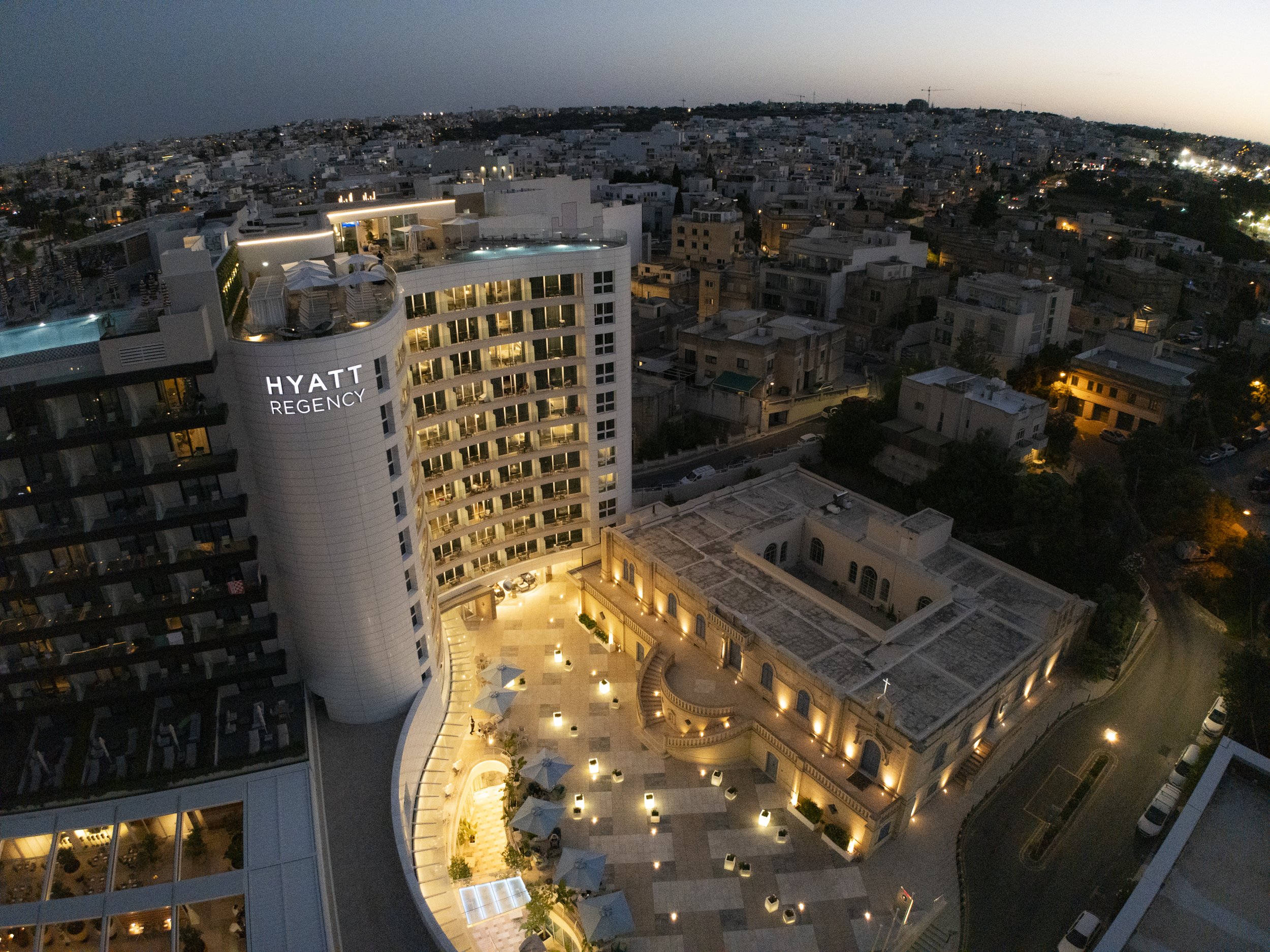 An aerial view at dusk of the Hyatt Regency hotel with illuminated balconies, a pool area on the rooftop, and an adjacent historic church with exterior lighting, surrounded by a dense cityscape.