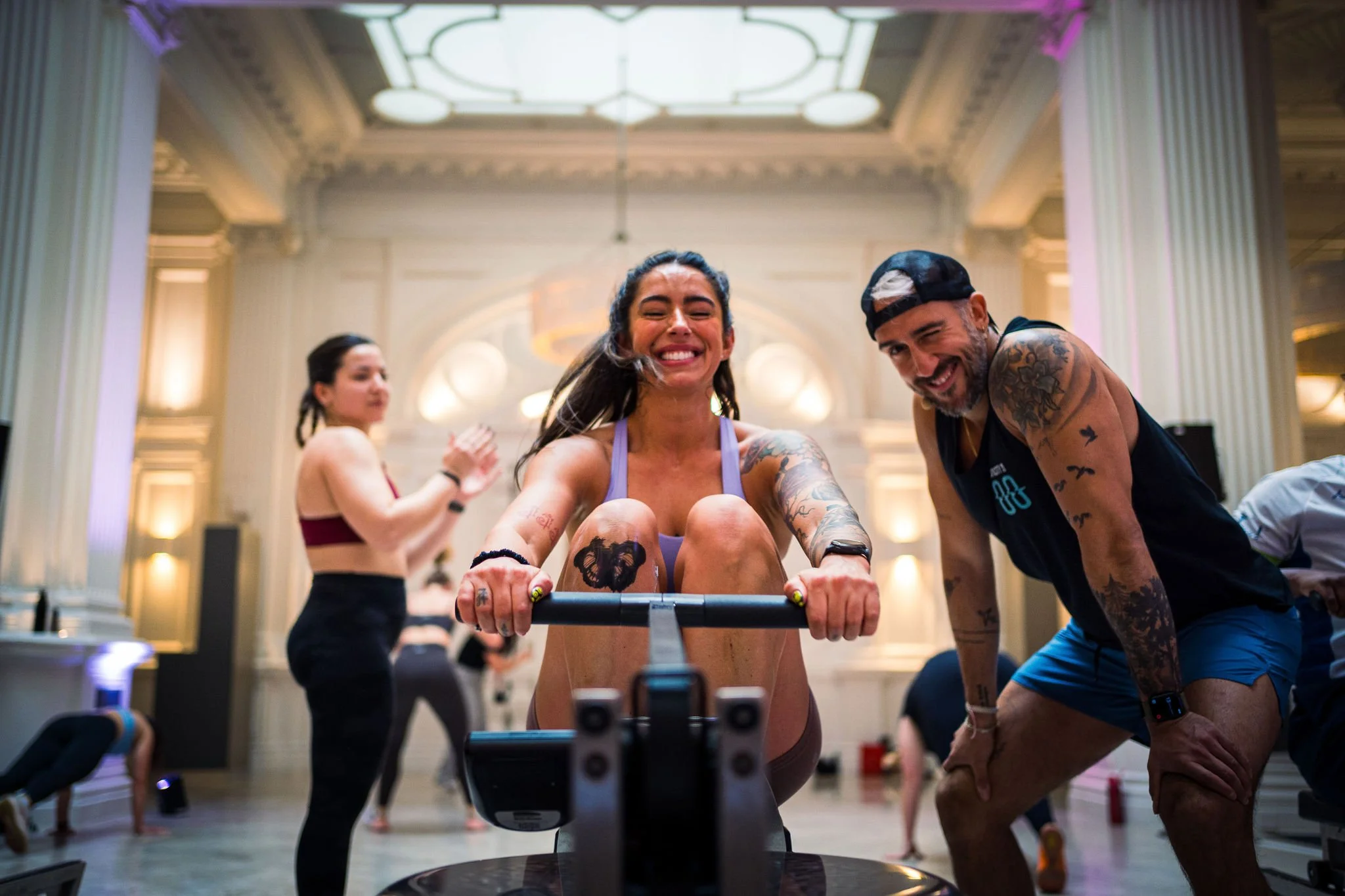 A woman smiling while using a rowing machine at an indoor fitness event, with a man in athletic wear beside her, and other participants in the background in a large, elegant room.