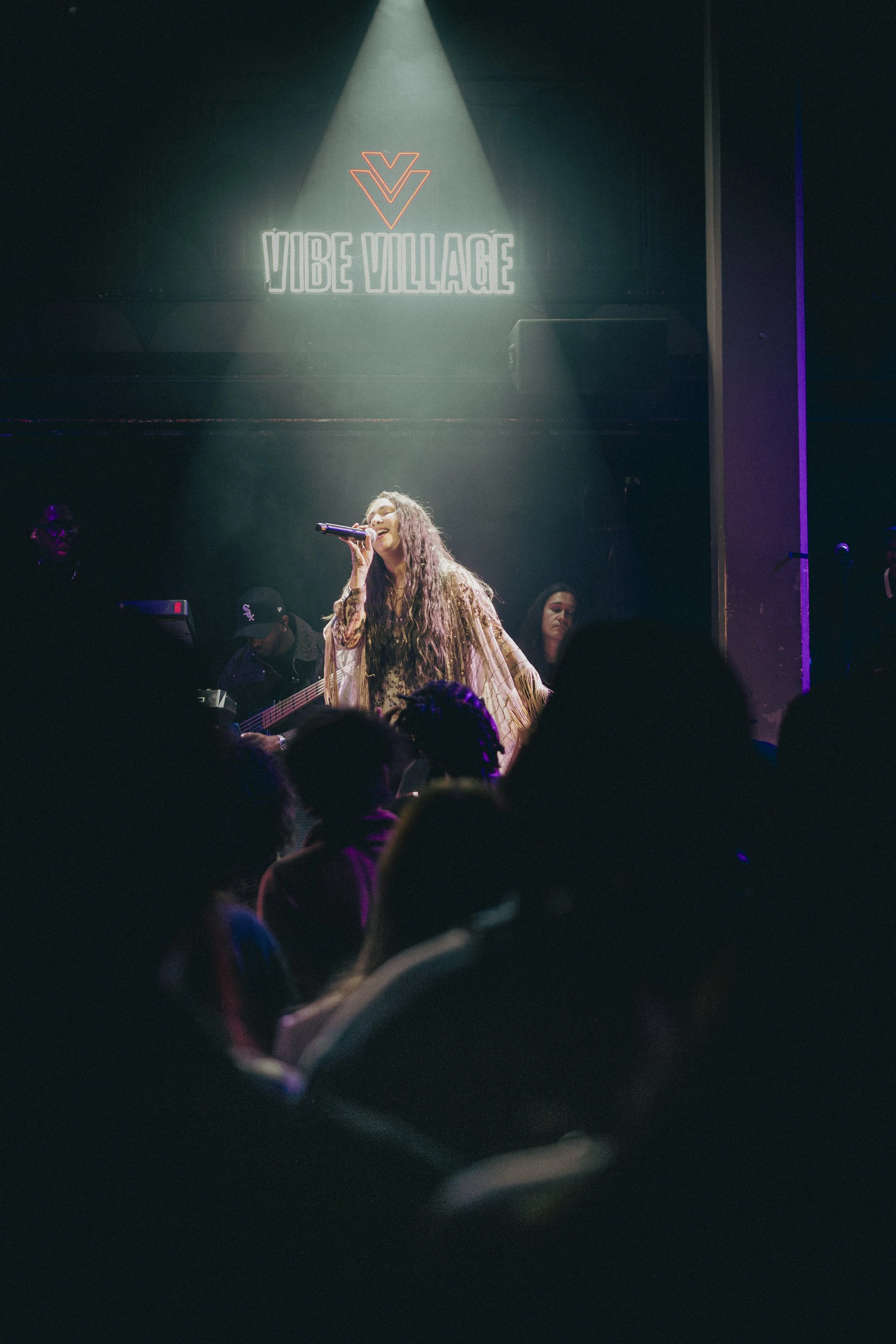 A woman singing into a microphone on stage with a band playing behind her, at Vibe Village music venue.