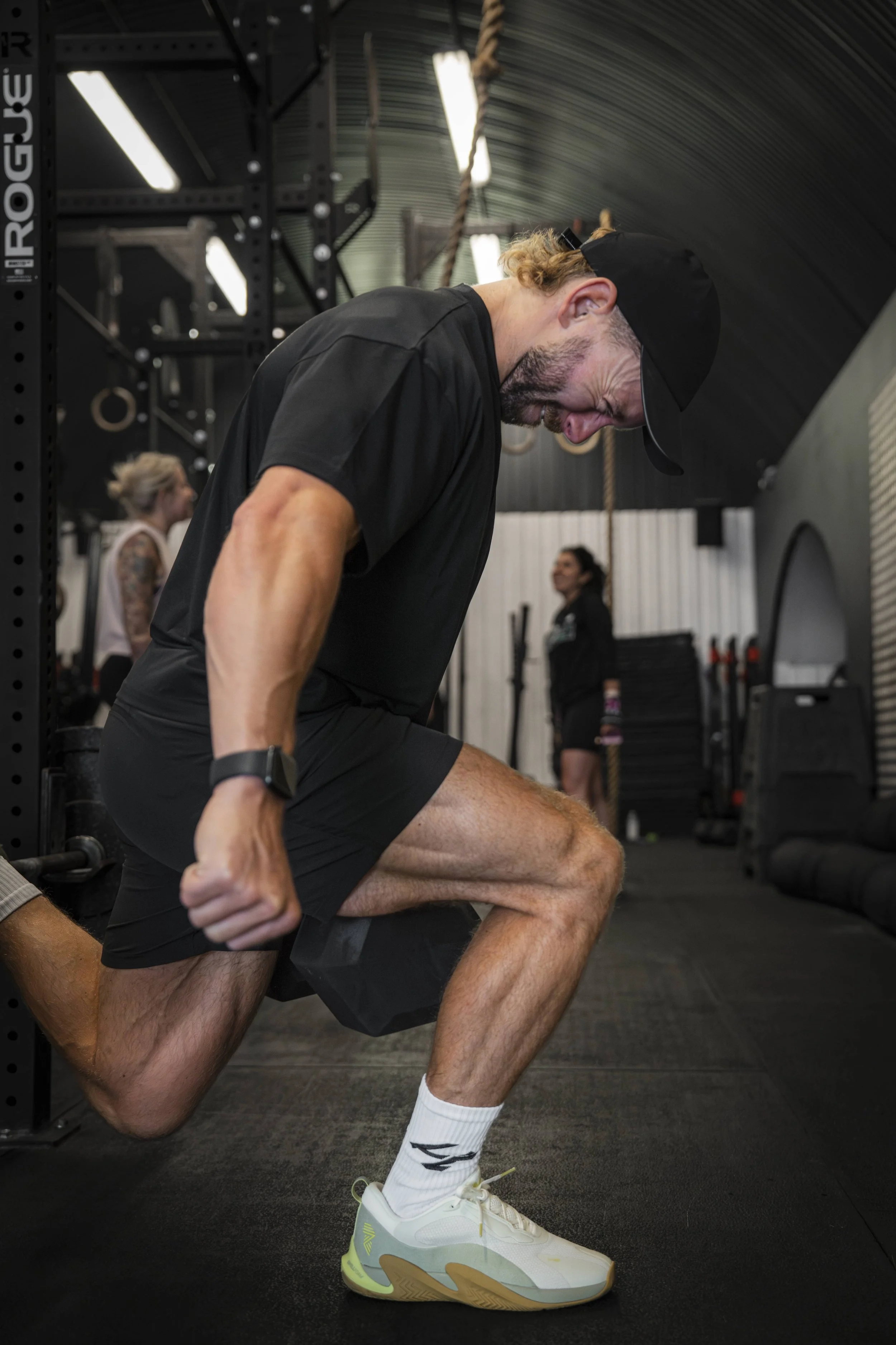 A man in black athletic clothing, cap, and white sneakers performs a pistol squat exercise in a gym, with a focused expression.