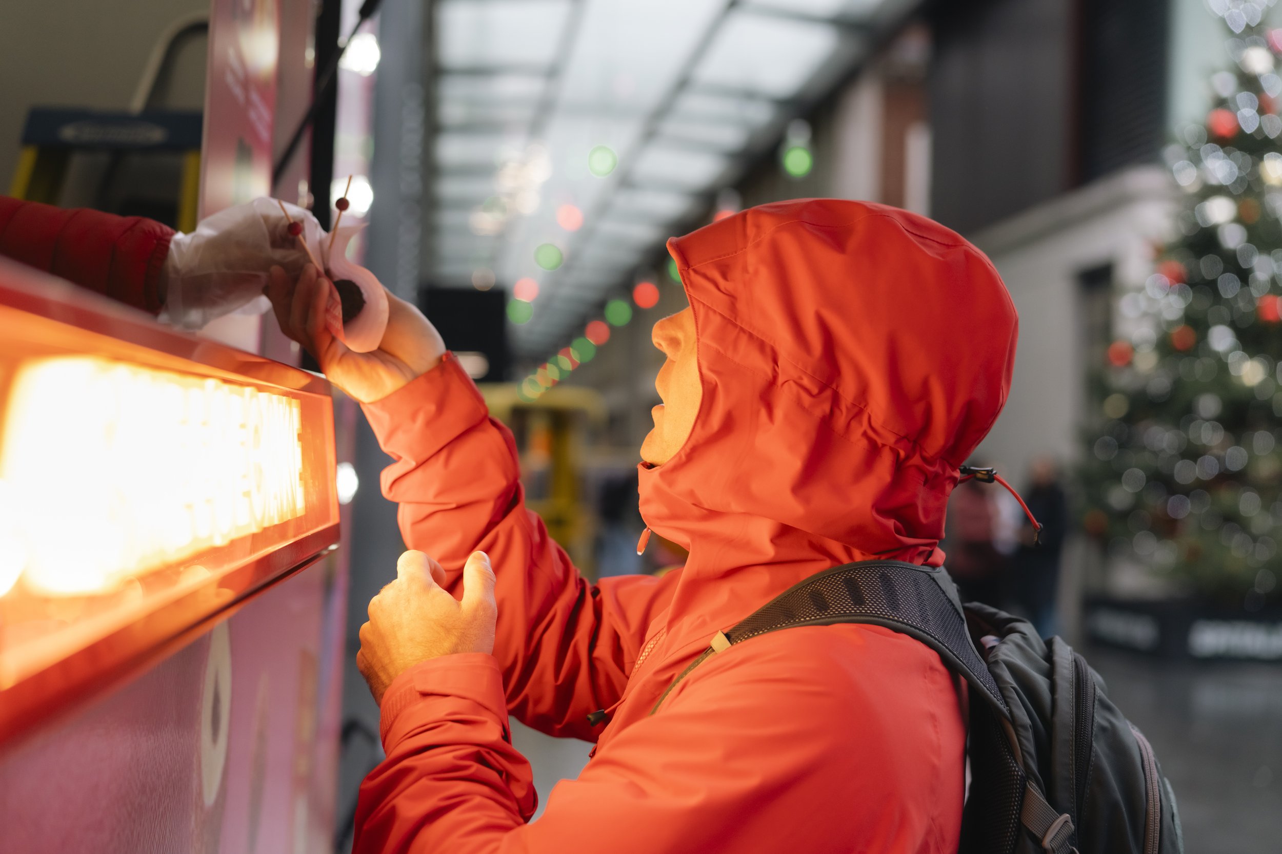 A person in an orange hooded jacket and a backpack is placing a coin into a donation box, with blurred holiday lights and a Christmas tree in the background.