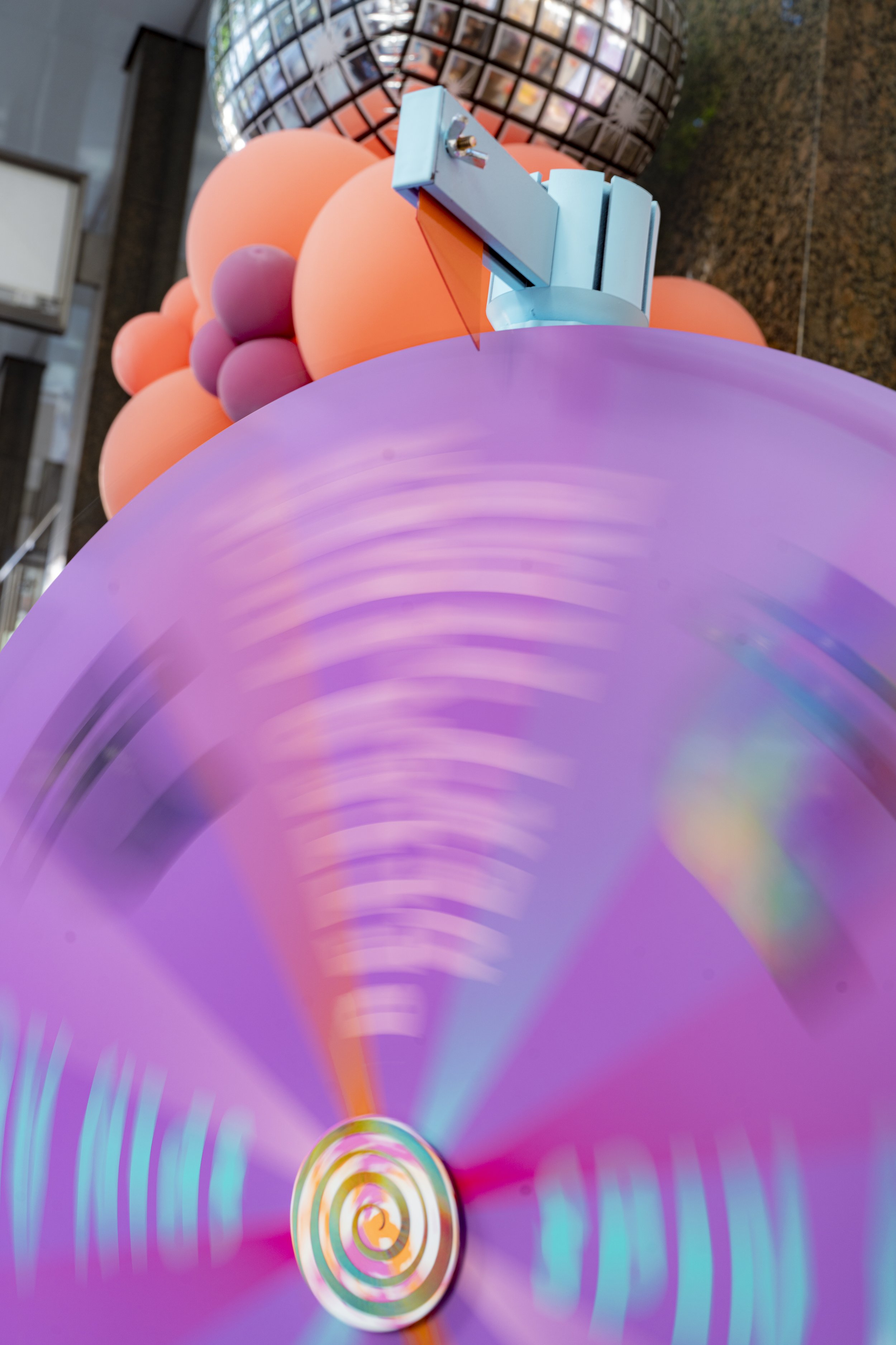 Close-up of an abstract sculpture with pink, purple, and orange spherical shapes and a metallic, blue geometric structure on top, with a purple background.
