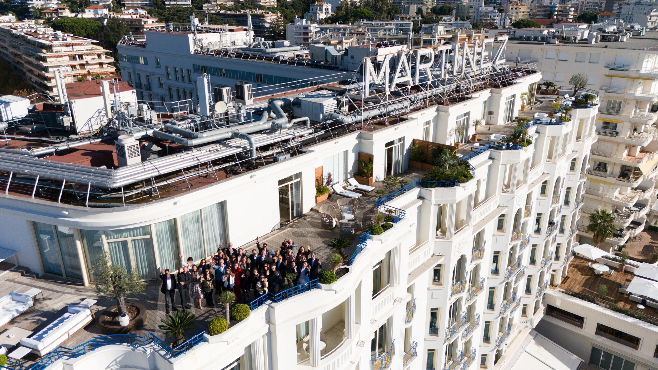 A group of people on a rooftop terrace of a tall white building with a 'Marina' sign on the roof, overlooking other buildings in an urban area.