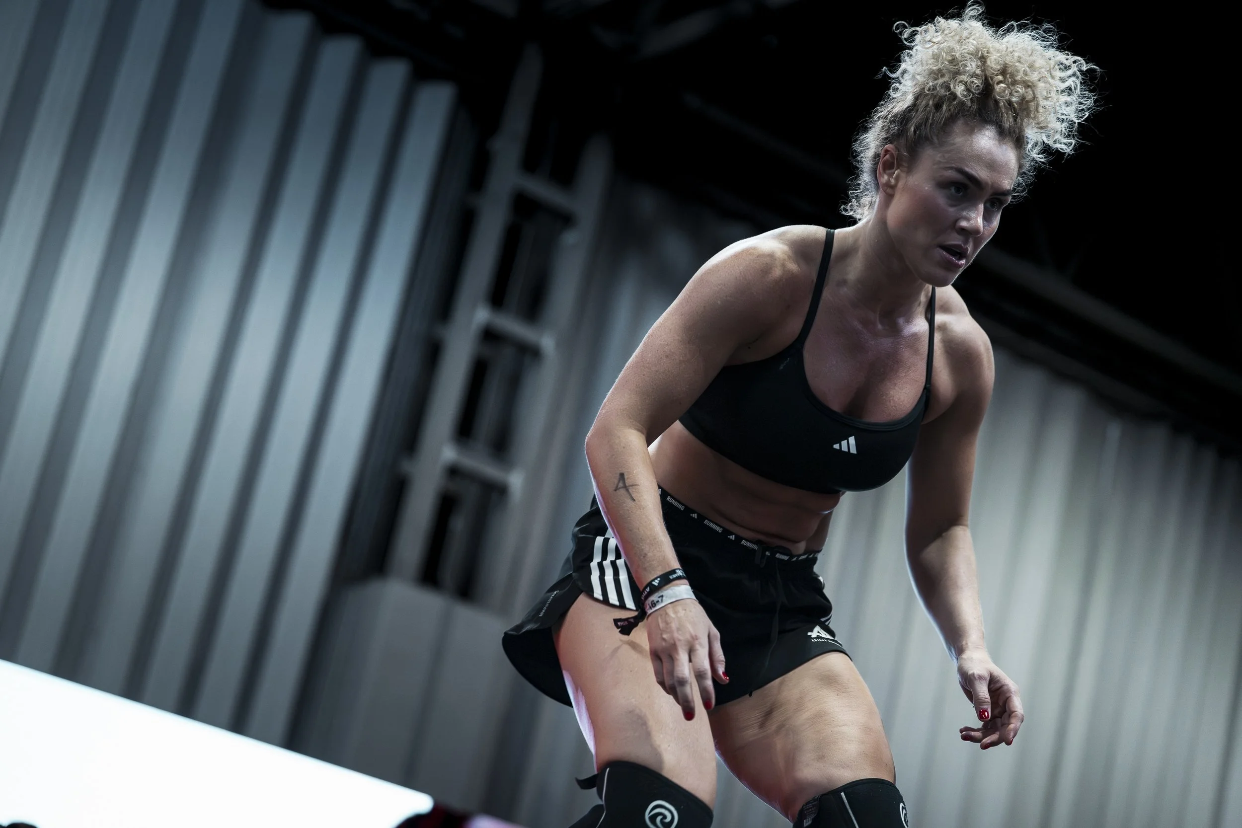 Female athlete with curly blonde hair in workout gear, looking exhausted and sweating, in an industrial gym setting.