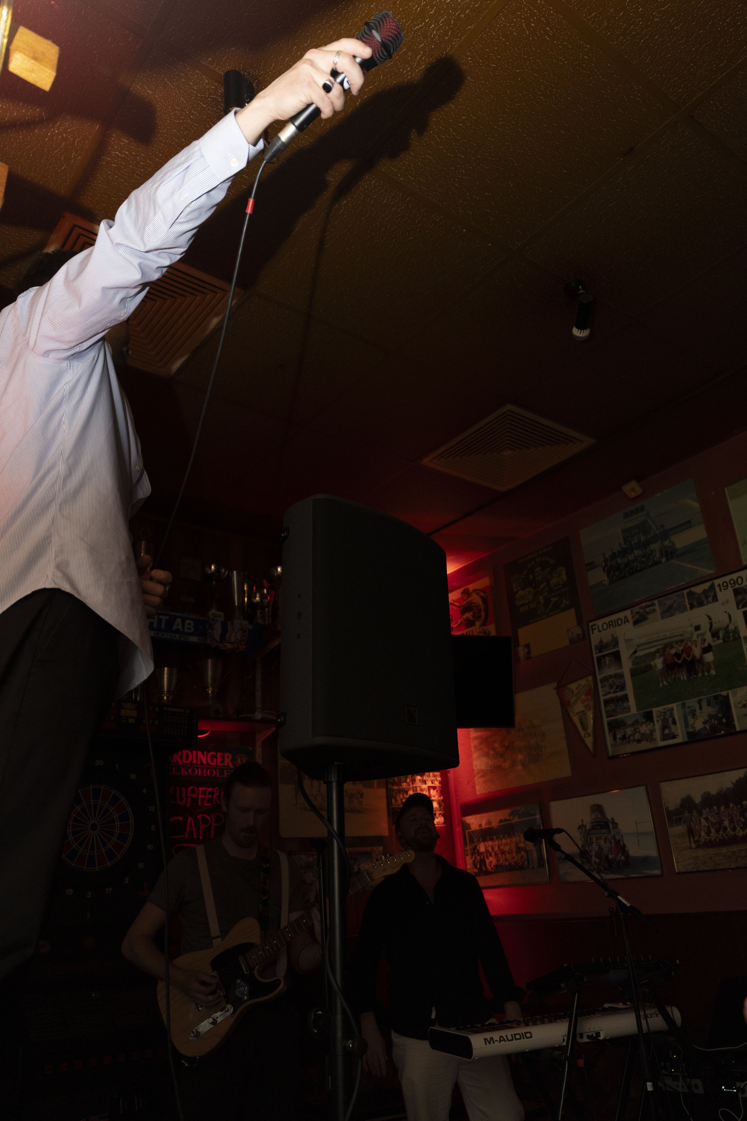 A person holding a microphone in a dimly lit bar or club with two musicians playing instruments in the background, surrounded by framed photos and memorabilia on the wall.