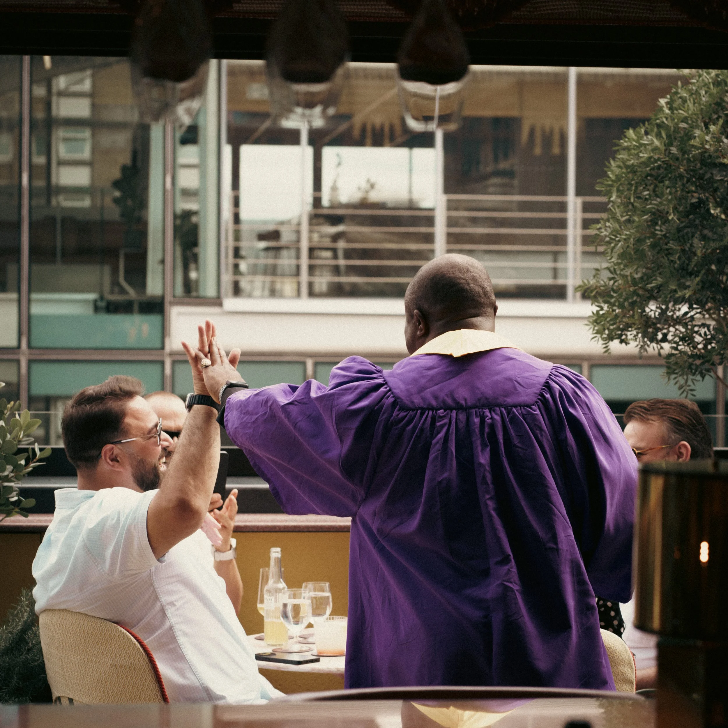 A man in a white shirt and glasses is giving a high-five to another man in a purple graduation gown at an outdoor celebration.