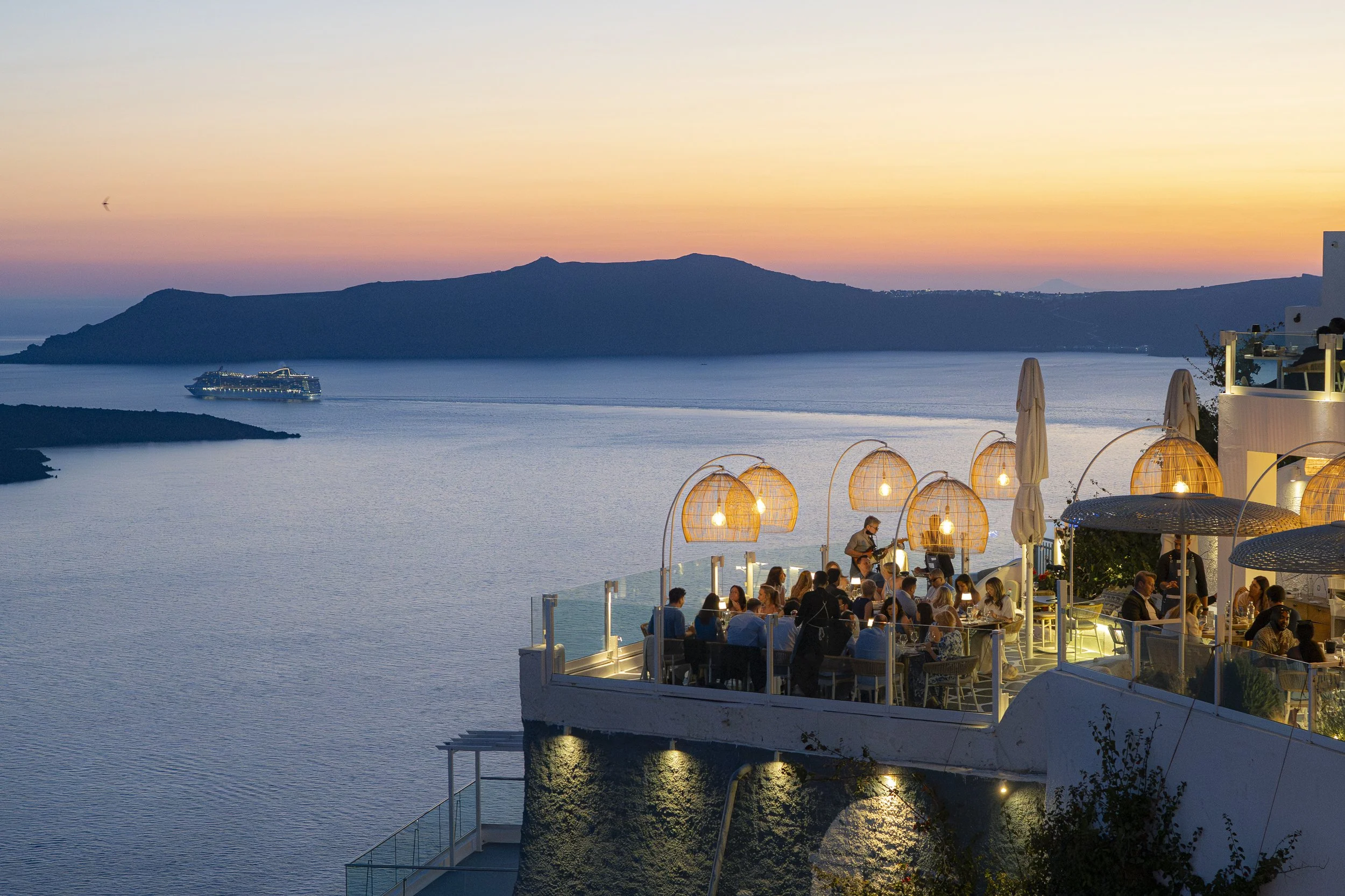 A seaside restaurant on a cliff during sunset, with people dining under hanging lamps, overlooking a calm sea with a cruise ship in the distance and a mountainous island in the background.